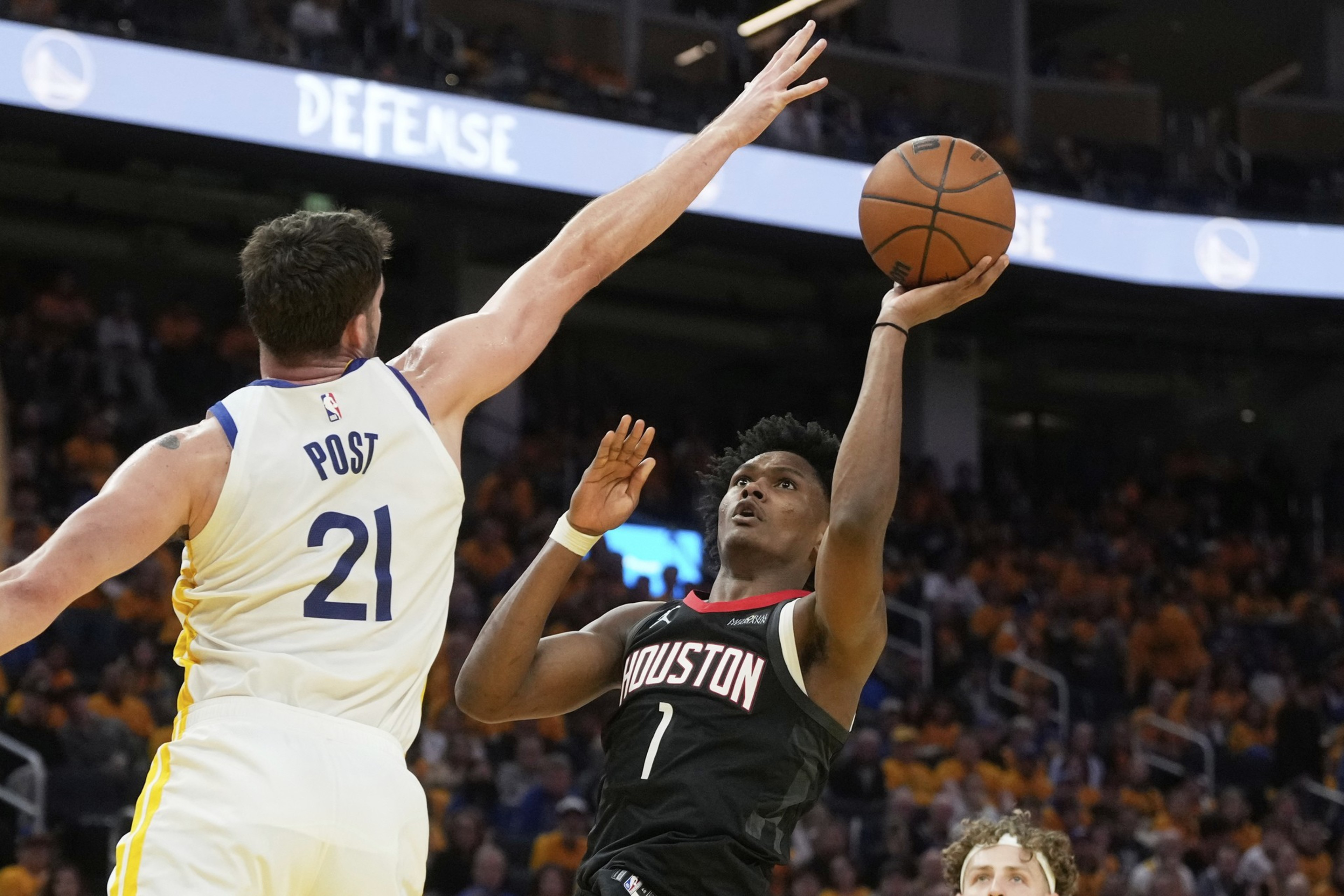 A basketball player in a black Houston jersey attempts a layup while a player in a white jersey labeled &quot;POST 21&quot; tries to block him. The crowd watches intensely.