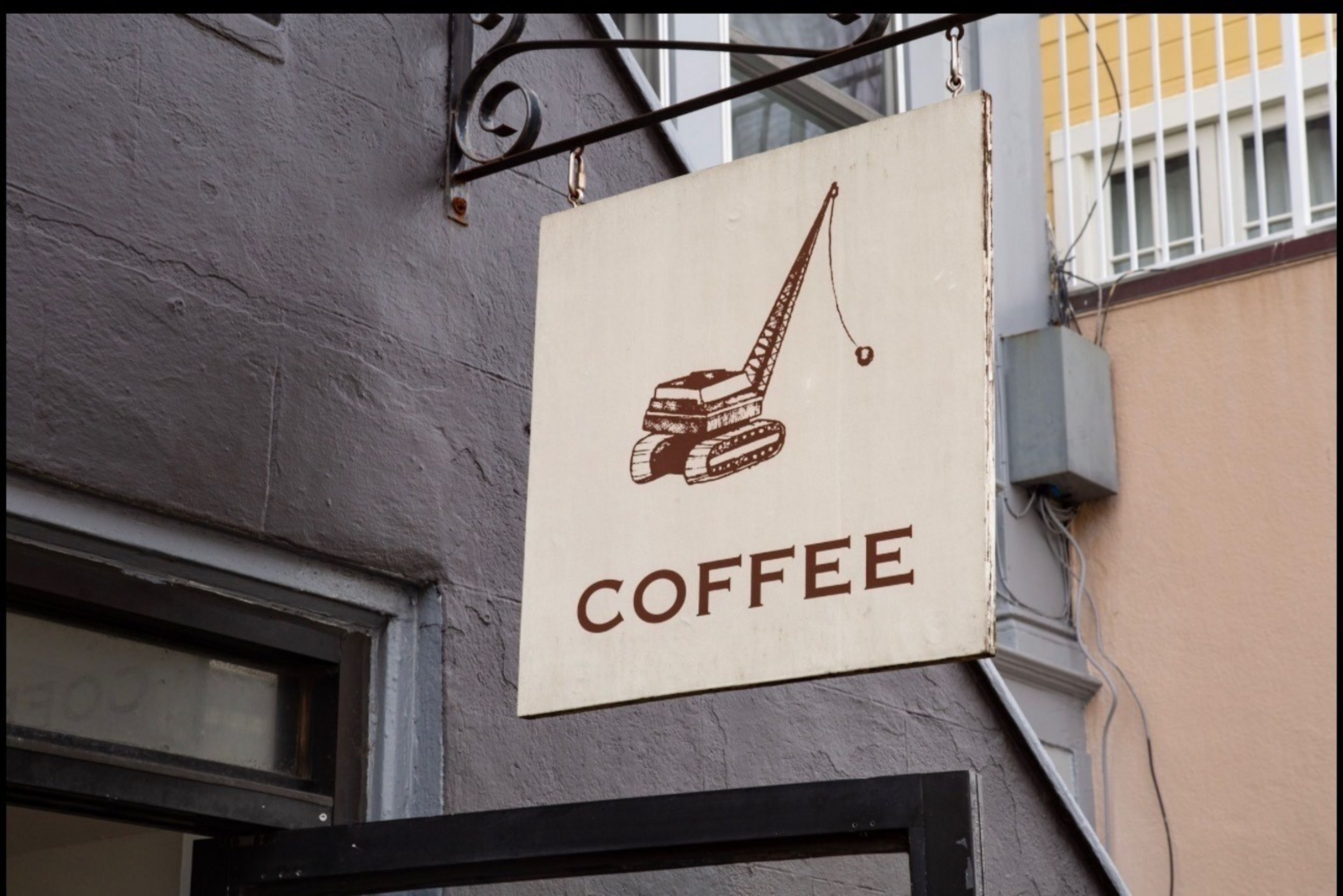 A rectangular coffee shop sign hangs on a metal bracket, featuring a brown illustration of a crane and the word "COFFEE" beneath it on a beige background.