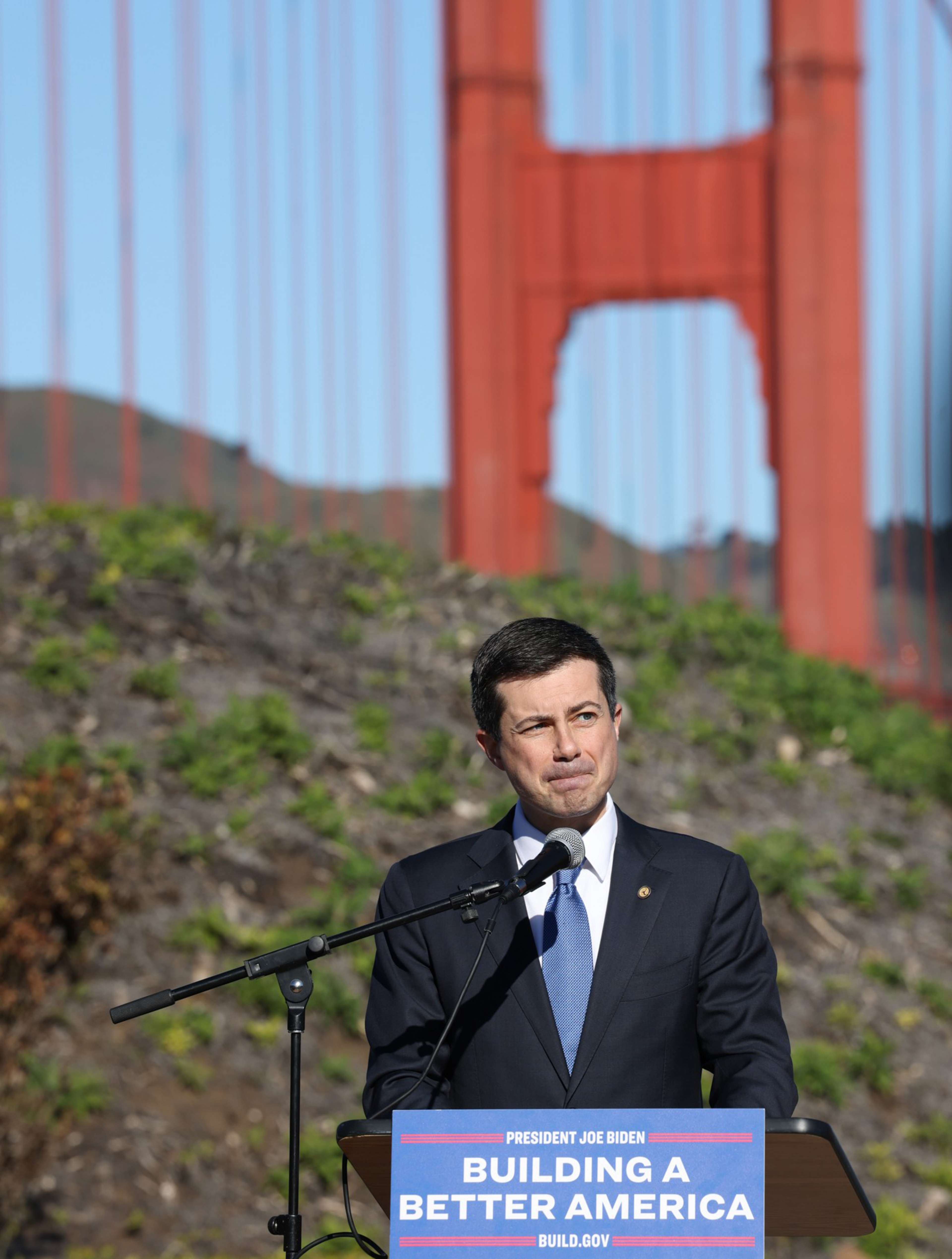 A man in a suit speaks at a podium with a "Building a Better America" sign, with the Golden Gate Bridge and greenery in the background.