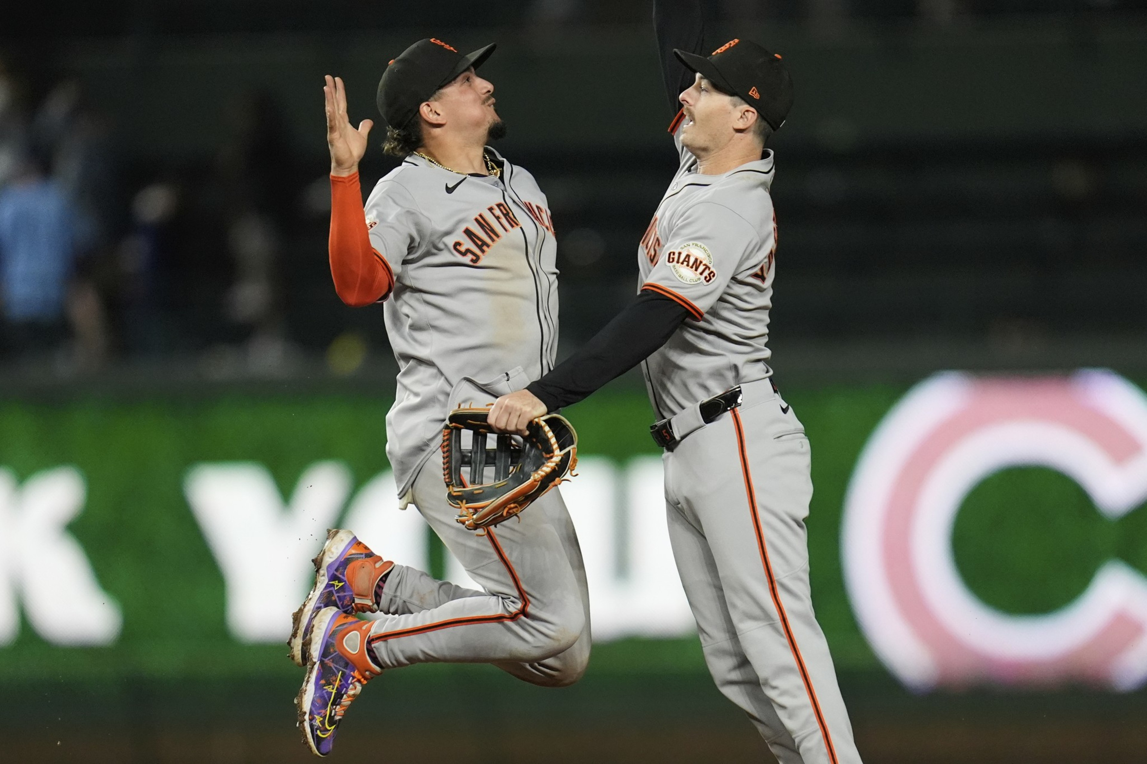 Two baseball players in gray uniforms jump and high-five in celebration on the field. One player holds a glove, and both are wearing black caps with orange details.