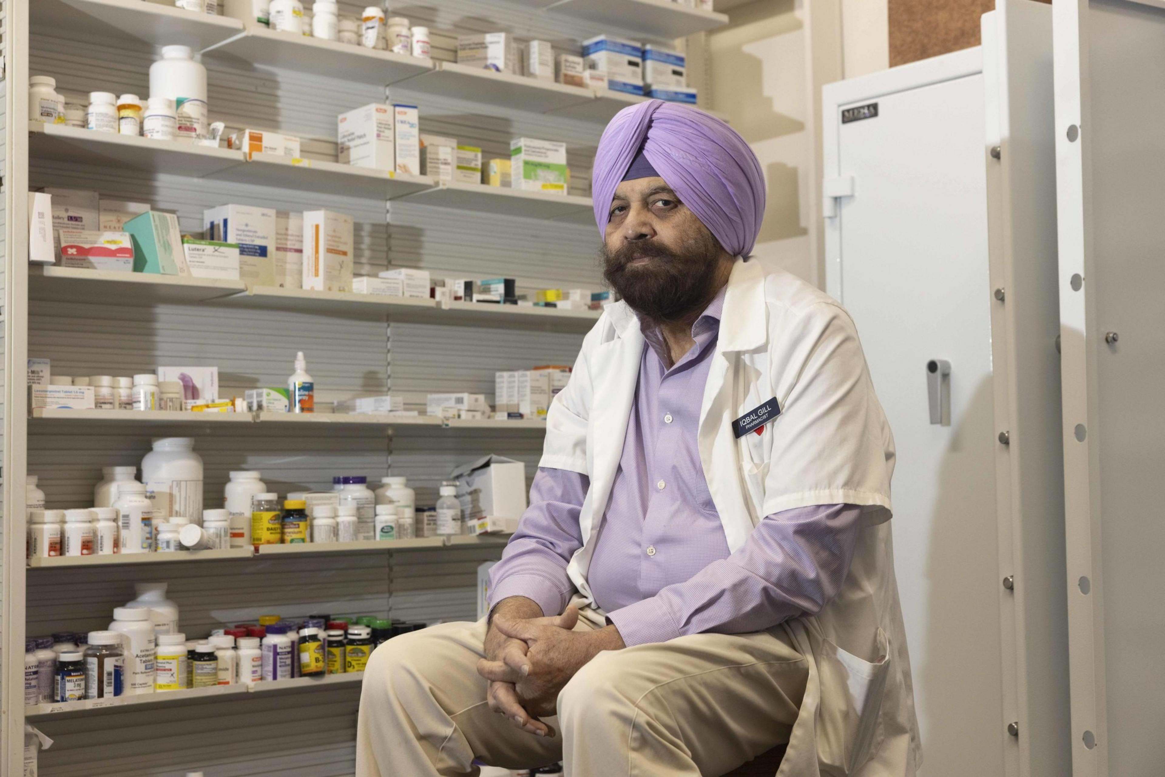 A man in a purple turban and white lab coat sits in a pharmacy, surrounded by shelves filled with various medicine bottles and boxes.