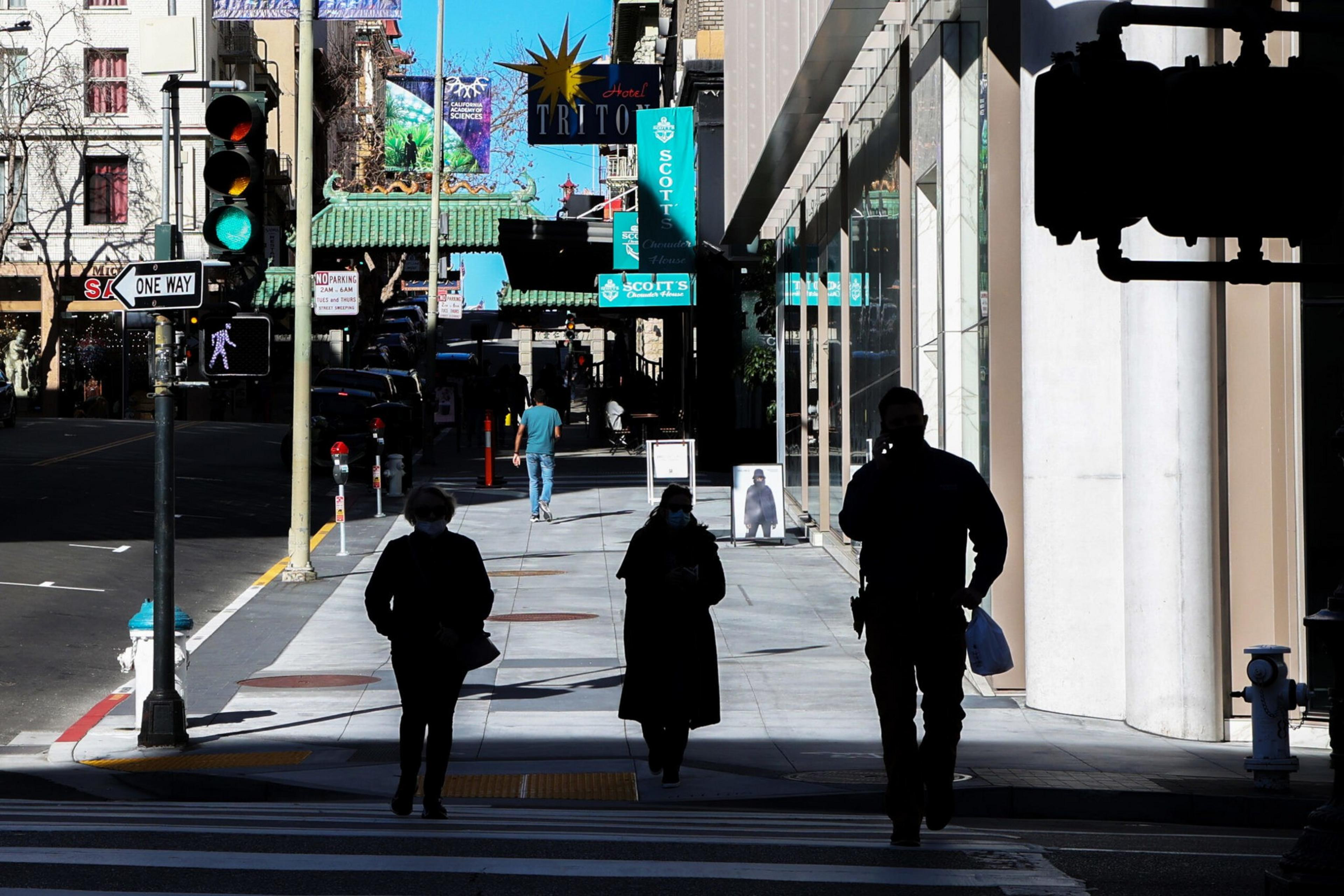 Three people walking on a city sidewalk with shadows, a "one way" sign, traffic light, and storefronts visible under a bright blue sky.