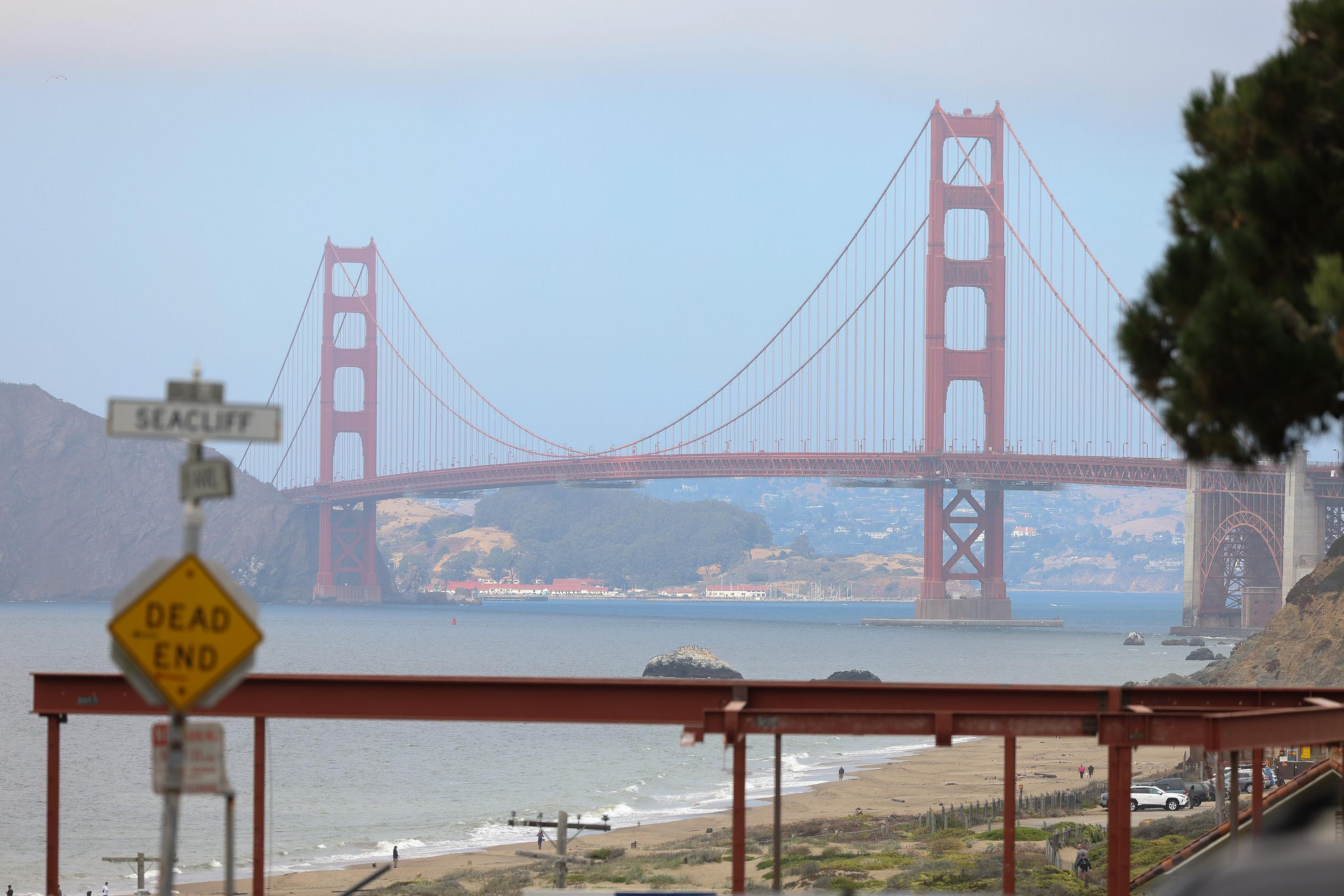 The image shows the Golden Gate Bridge spanning across the water with a clear sky. In the foreground, there's a &quot;Dead End&quot; sign and a beach with a few people.