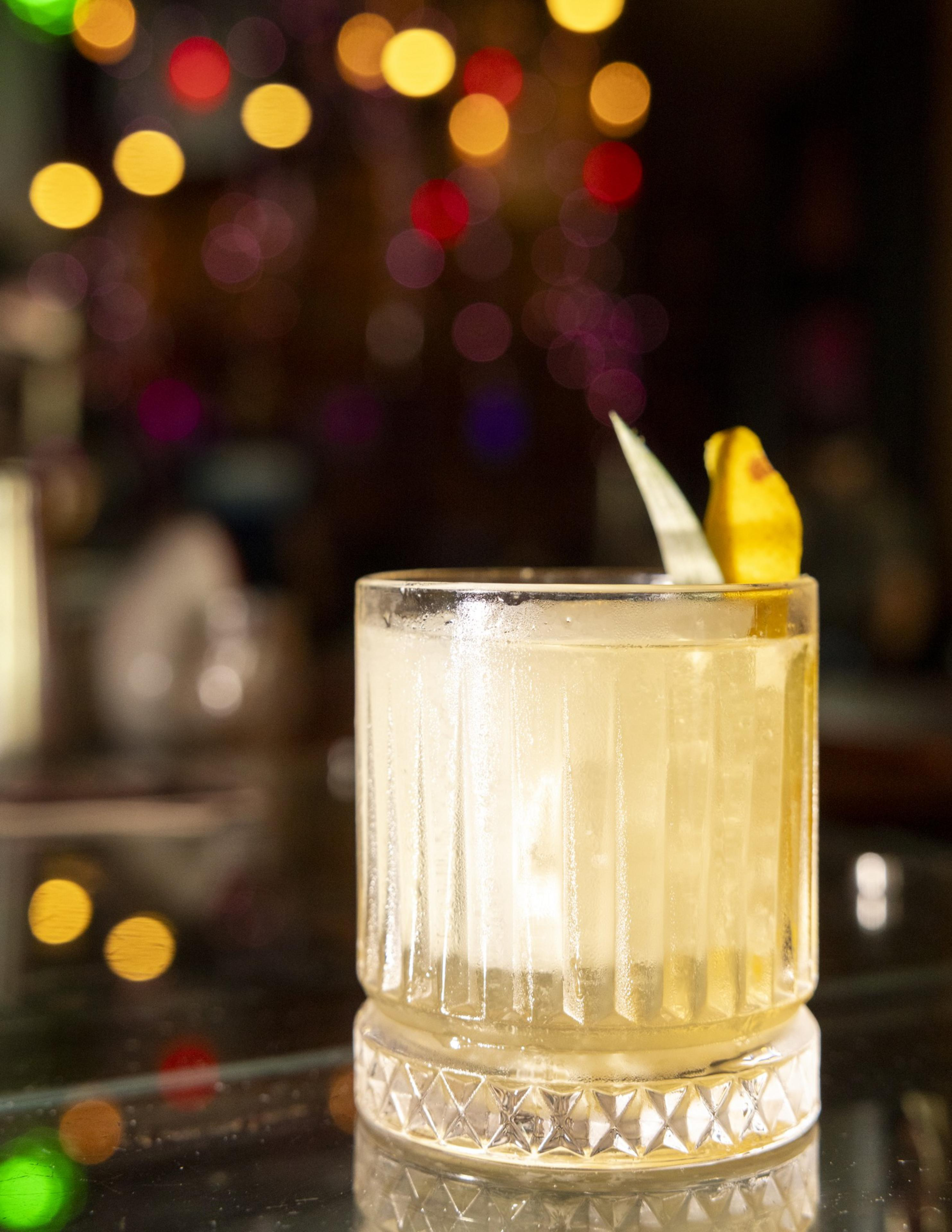 A frosty cocktail in a textured glass with a lemon twist garnish sits on a reflective surface, with colorful out-of-focus lights in the background.