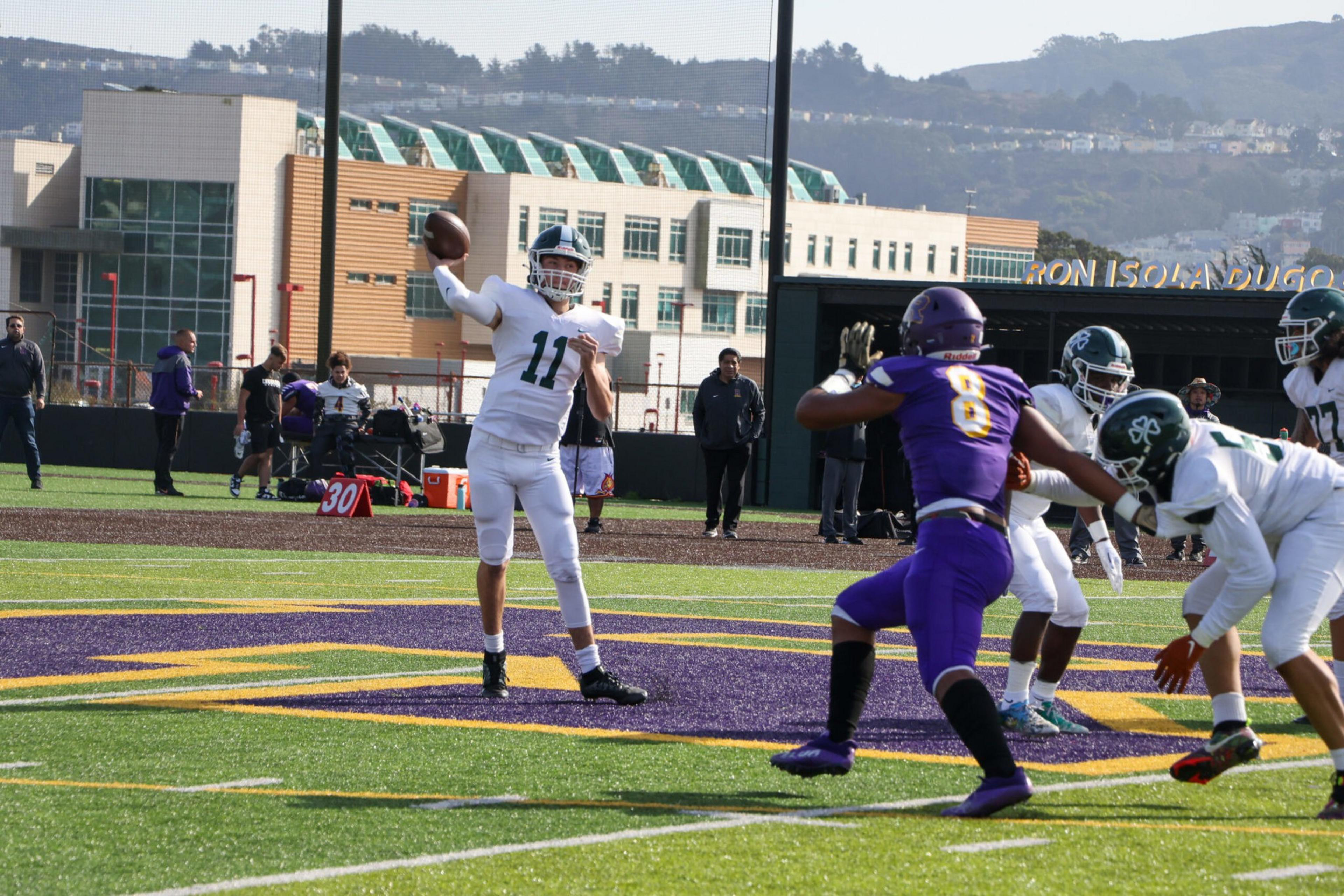 A football quarterback in white jersey number 11 is throwing the ball while a player in purple jersey number 8 moves to block him.