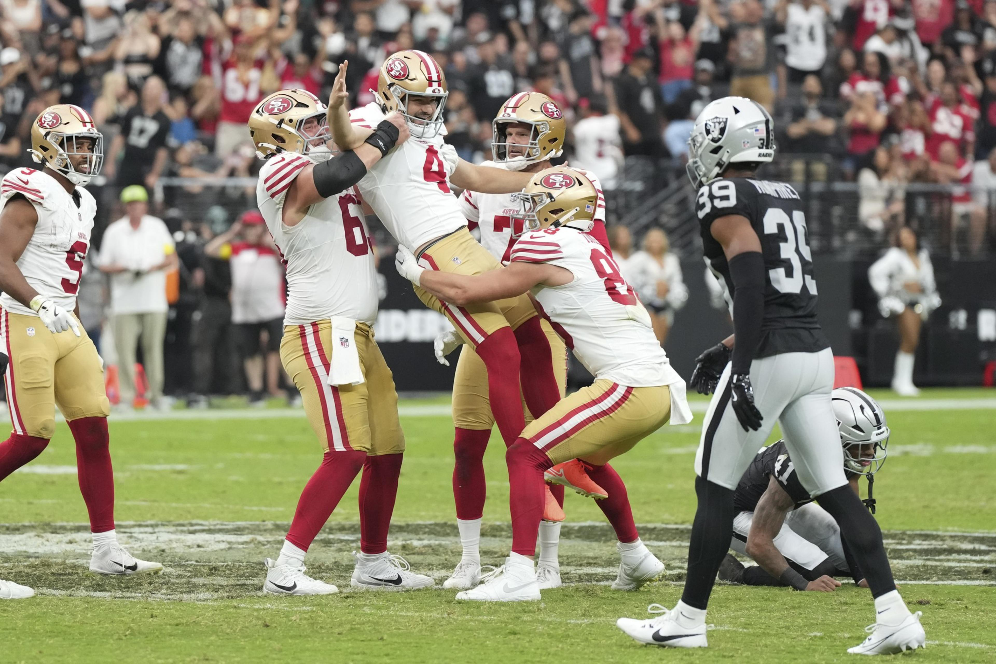 Four San Francisco 49ers players celebrate on the field while two Las Vegas Raiders players look on during a football game.