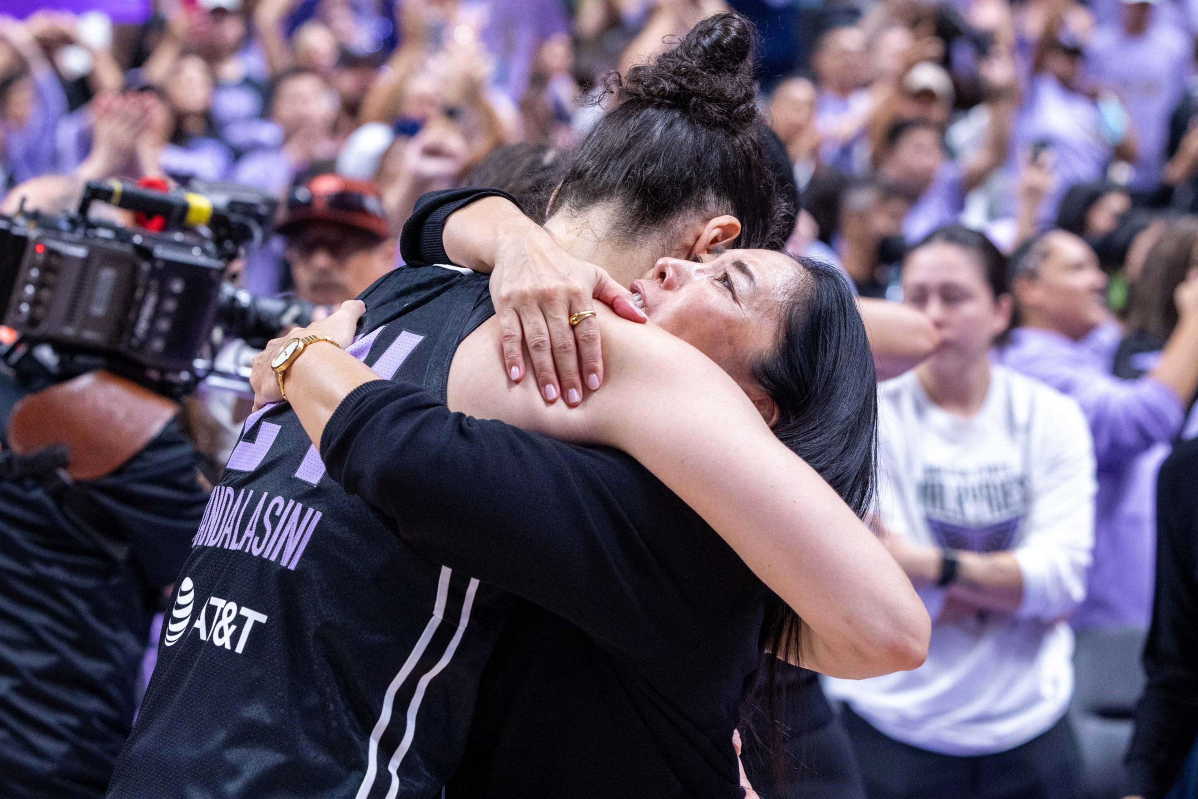 A basketball player and a woman share an emotional hug amidst a cheering crowd and a cameraman filming nearby.