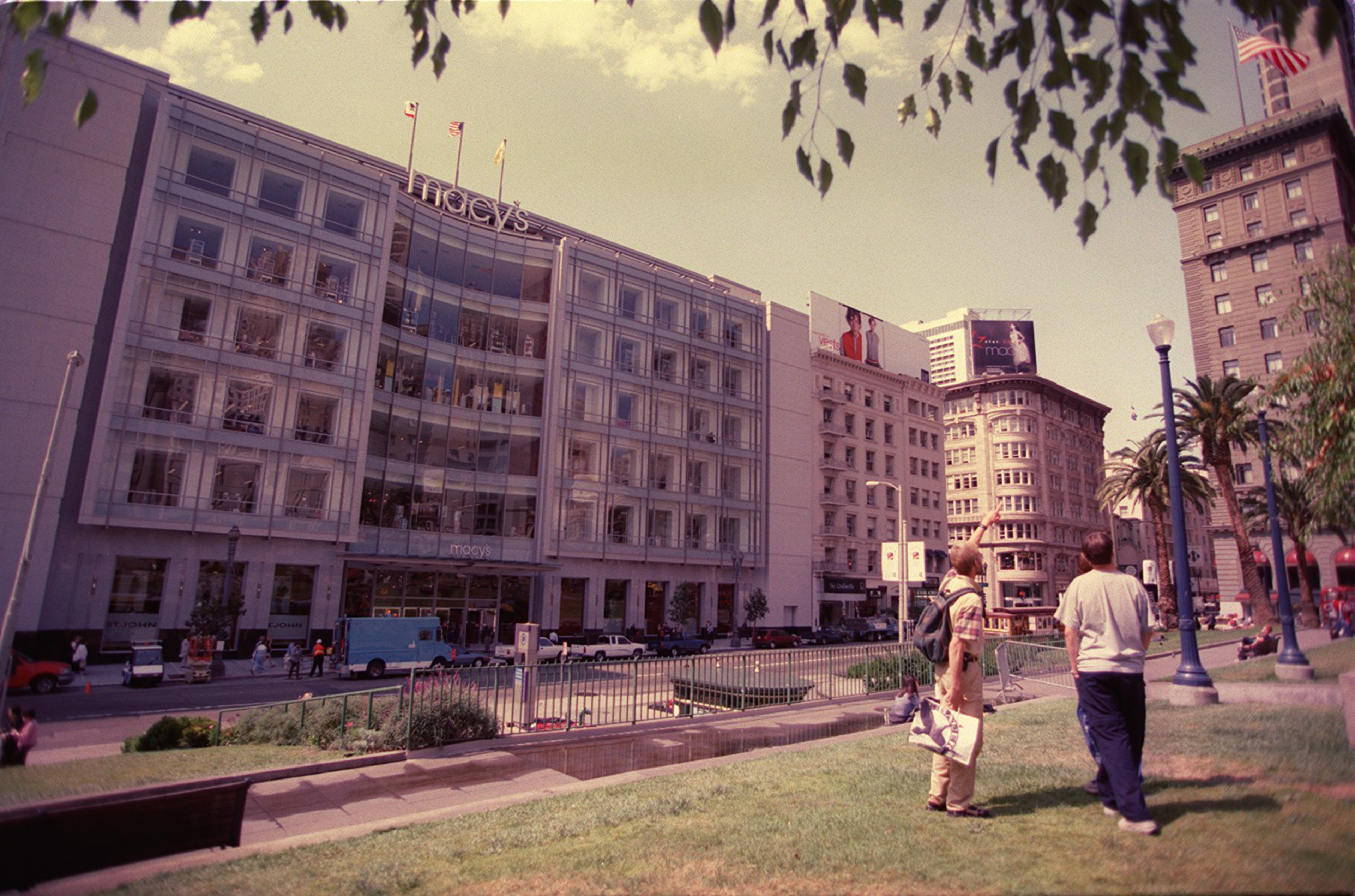 A sunny urban park with people, in front of a large Macy's store and city buildings.