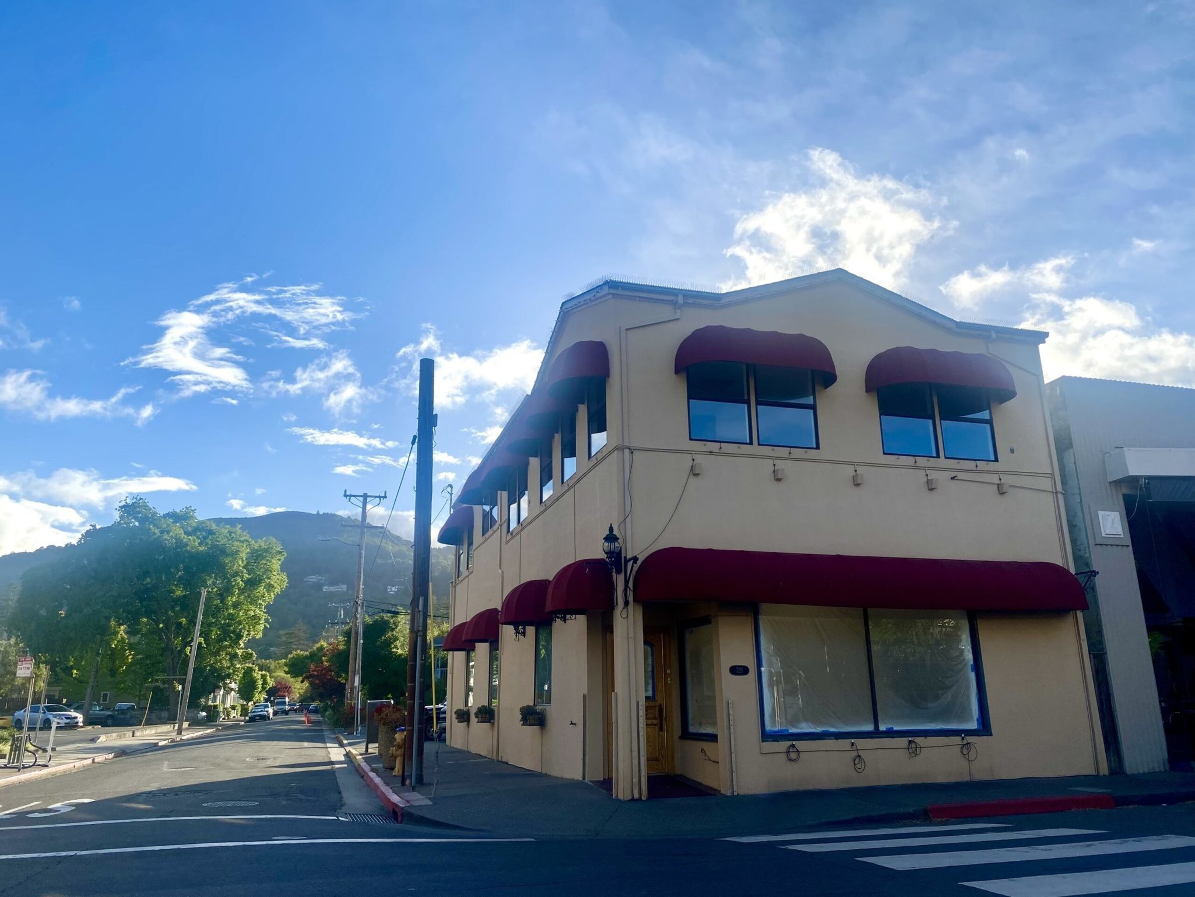 A two-story beige building with maroon awnings stands at a street corner under a bright blue sky with scattered clouds.
