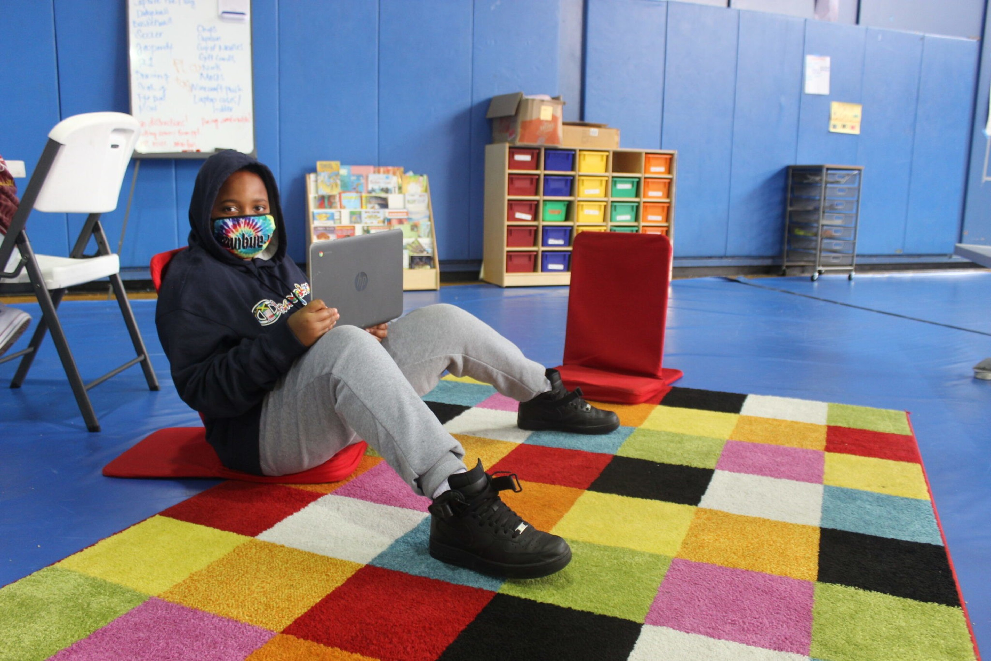 A child wearing a mask and hoodie sits on a red cushion on a colorful checkered rug, holding a laptop in a room with blue walls.