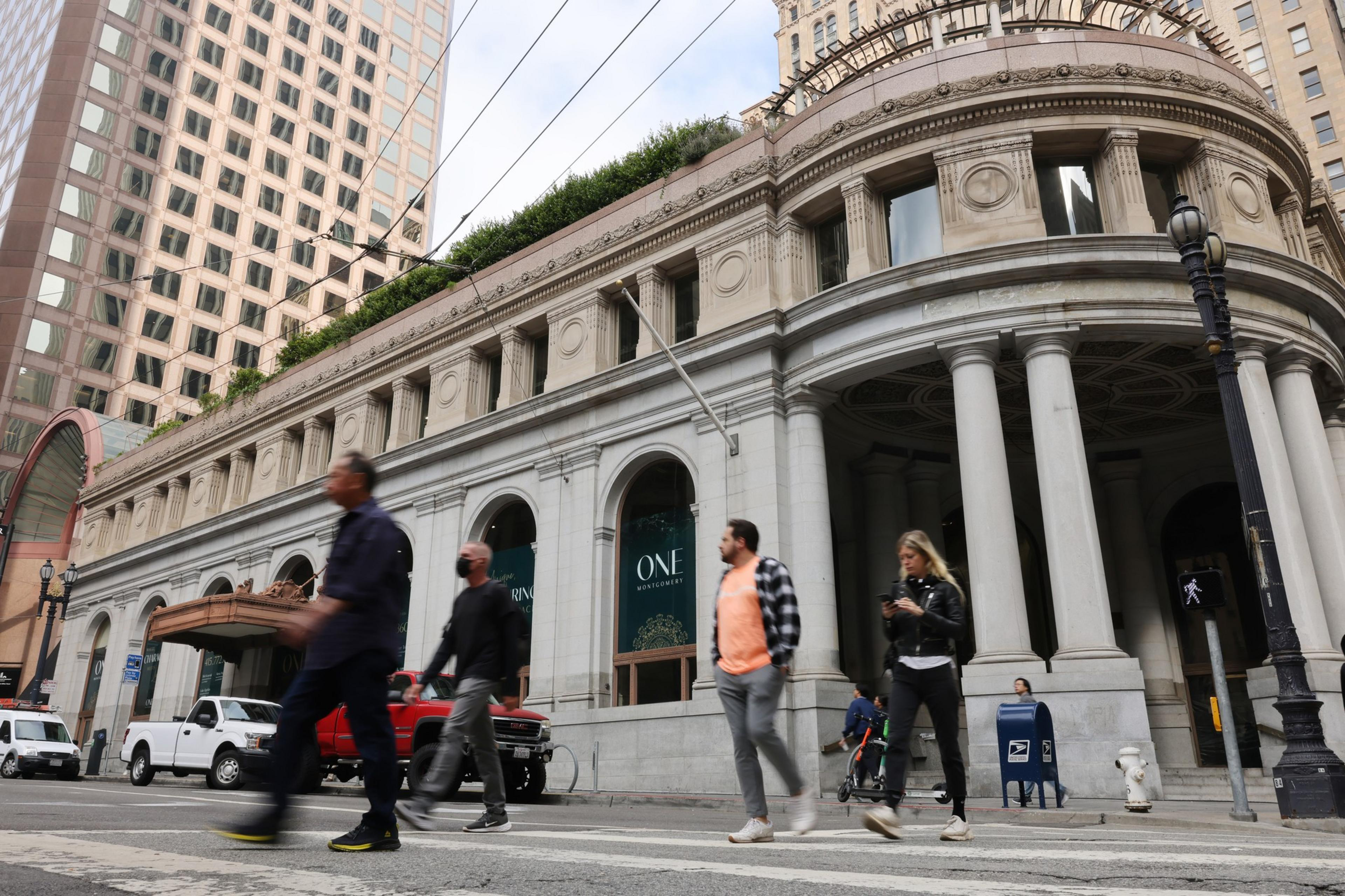 People cross a street in front of a grand, columned building. It's an urban scene with a mix of pedestrians and parked cars, under a partly cloudy sky.