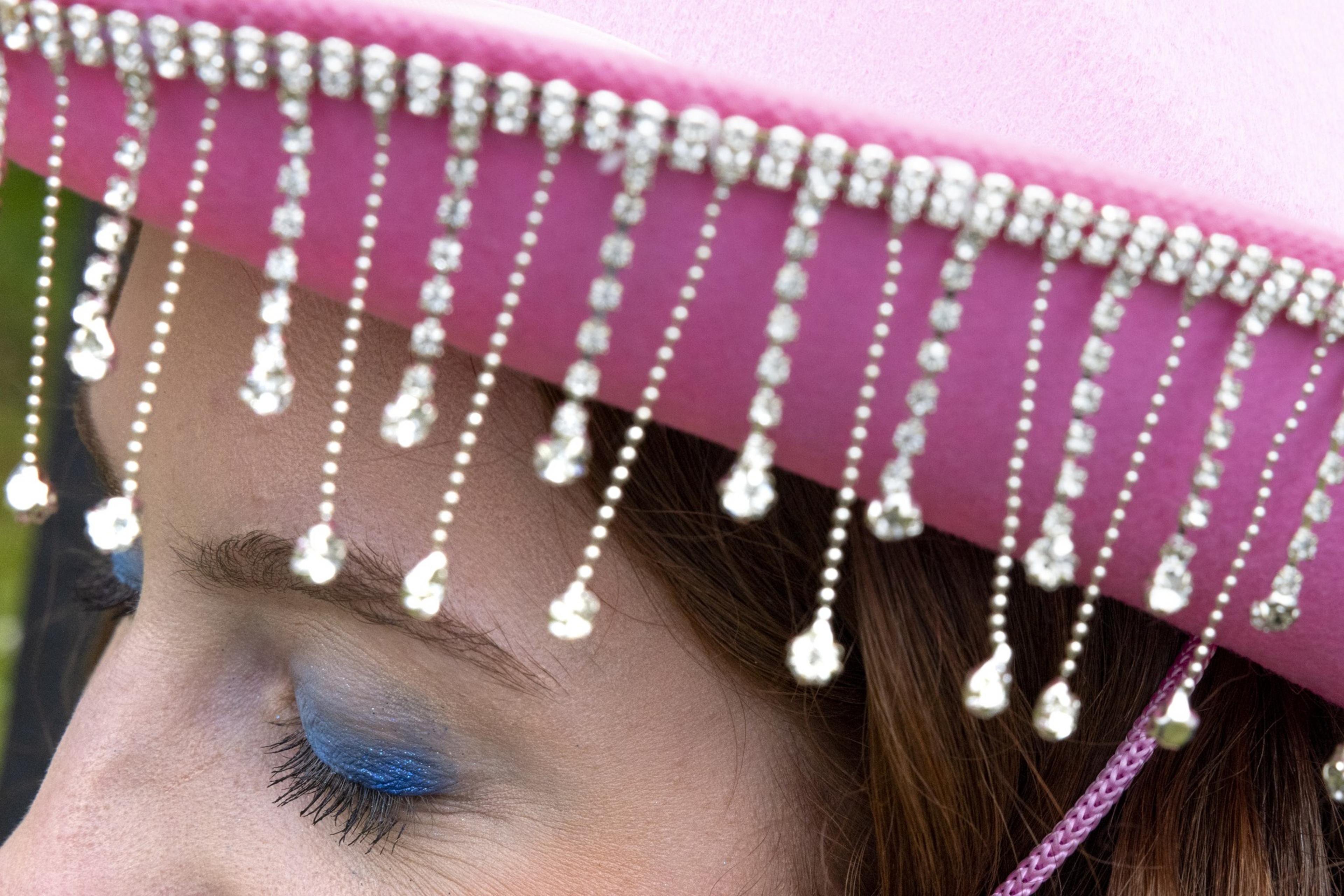 The image shows a person wearing a pink hat adorned with sparkling, dangling bead-like decorations. The person's eyes are closed and have blue eye shadow.