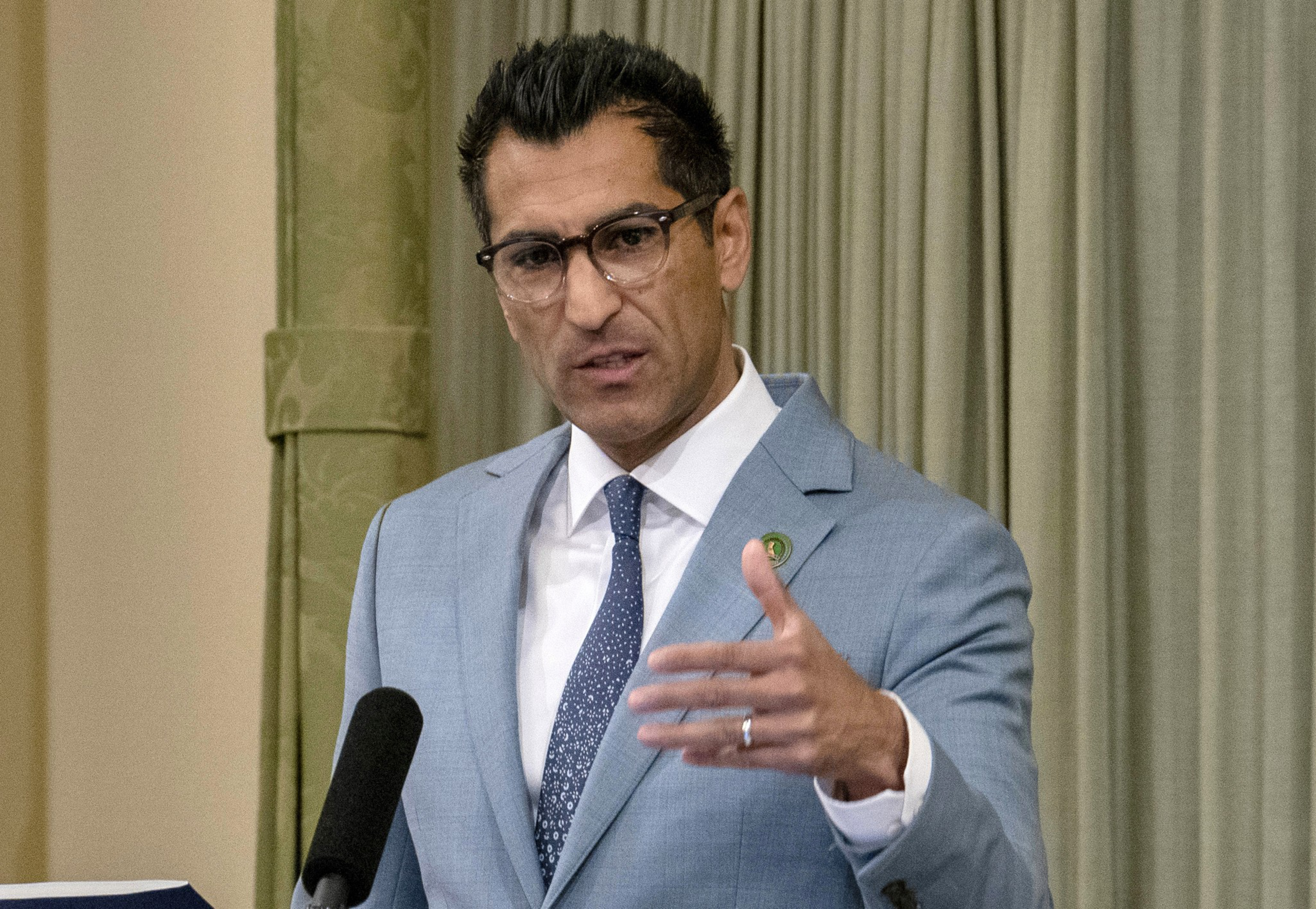 A man in a light blue suit and glasses gestures while speaking at a podium.