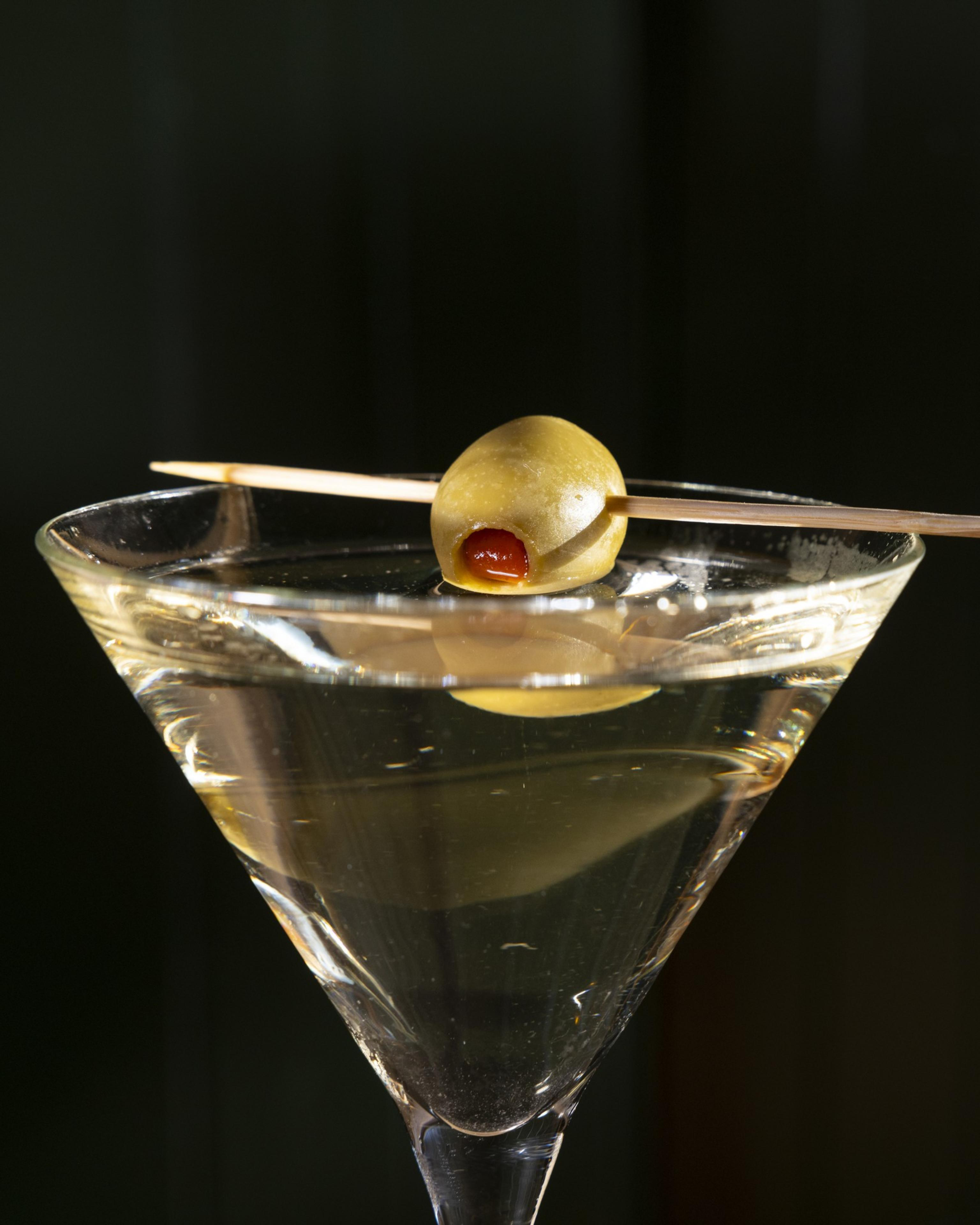 A clear martini glass holds a transparent drink, garnished with a single green olive on a cocktail stick placed across the rim.
