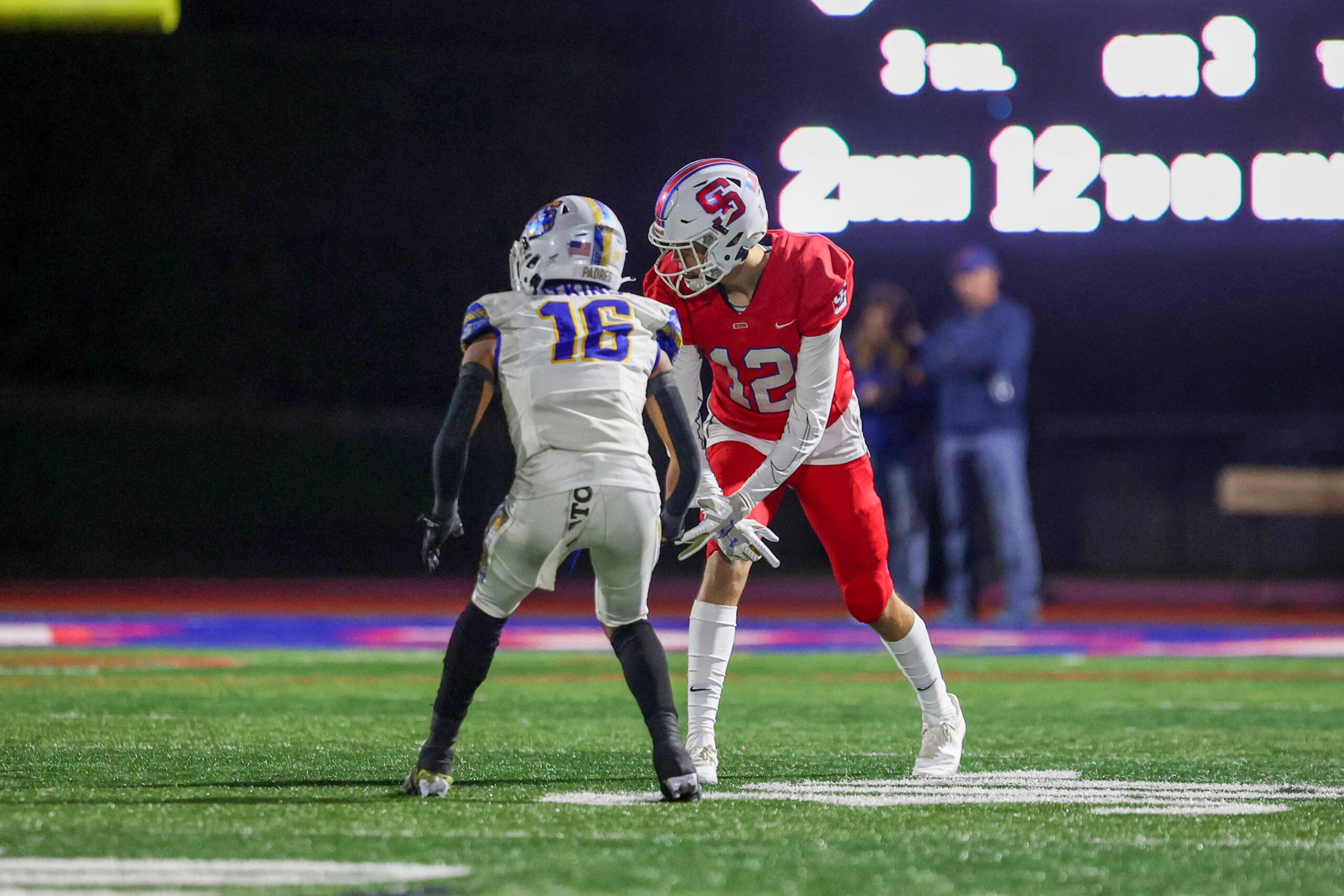 A football player in red prepares to catch the ball while facing a player in white, both standing on a green field with a scoreboard in the background.