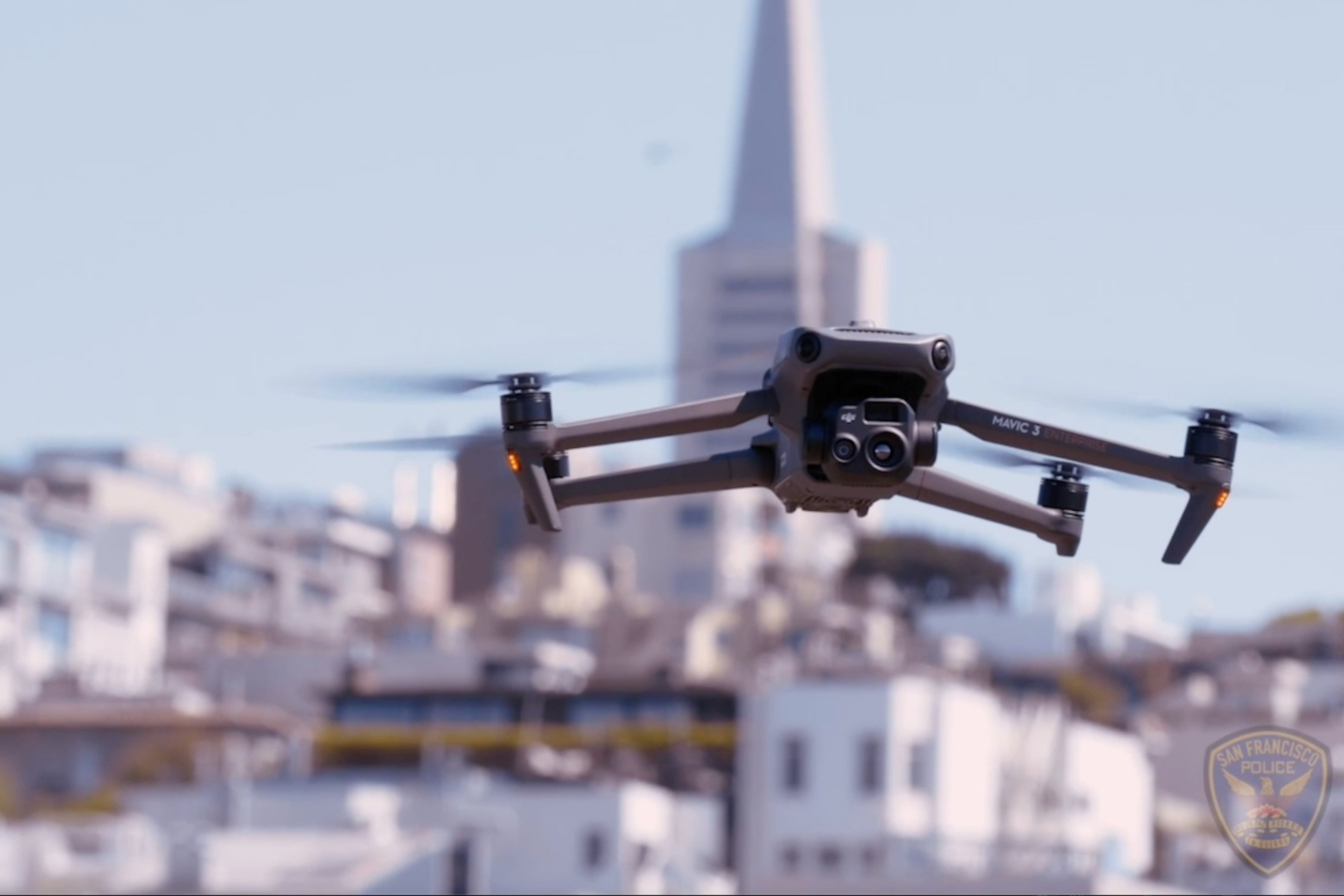 A DJI Mavic 3 drone hovers mid-air with blurred city buildings in the background and a small San Francisco Police badge in the corner.