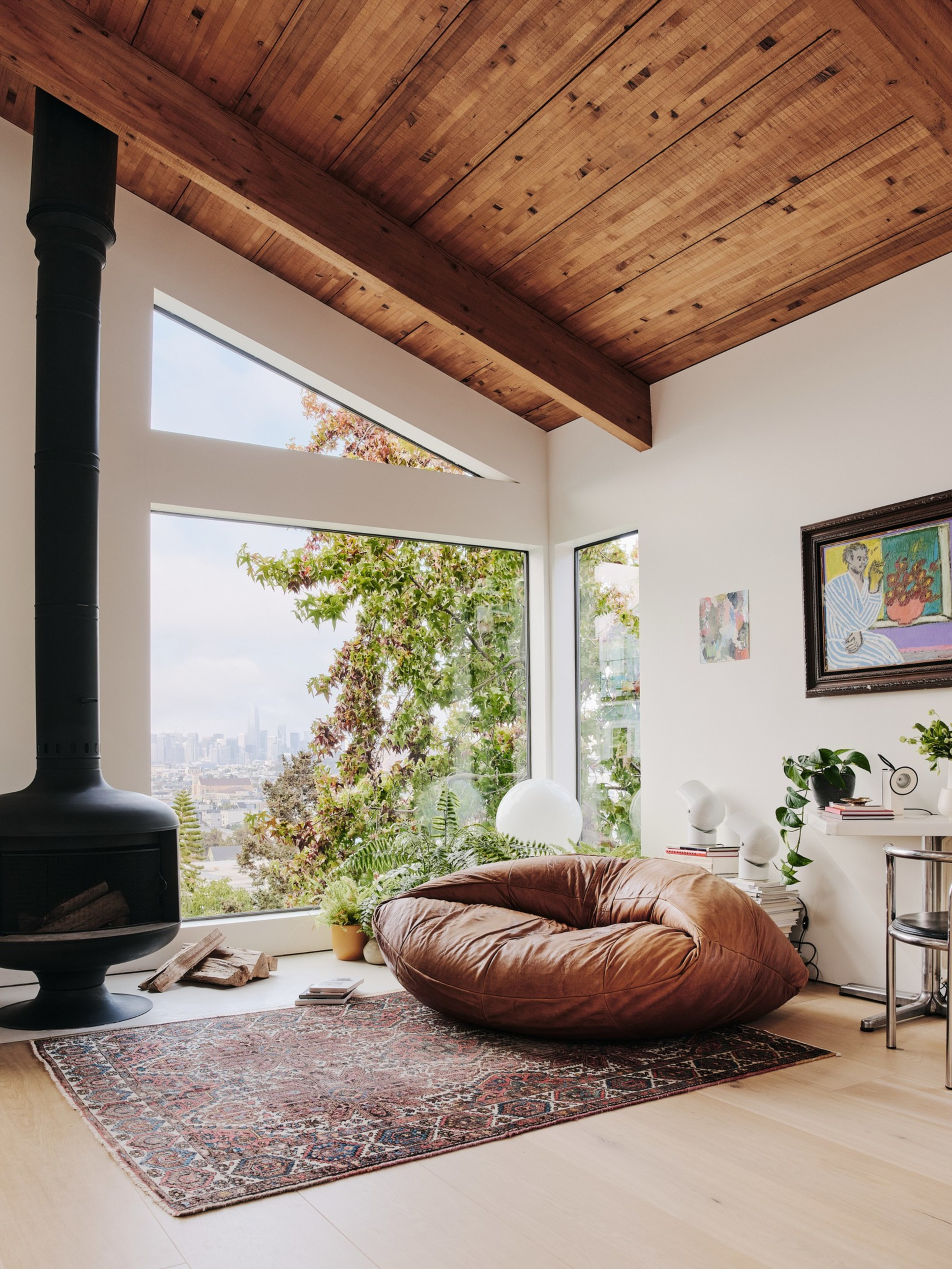 A cozy room features a leather bean bag on a patterned rug, a modern black fireplace, large windows with city views, plants, and art on the walls.