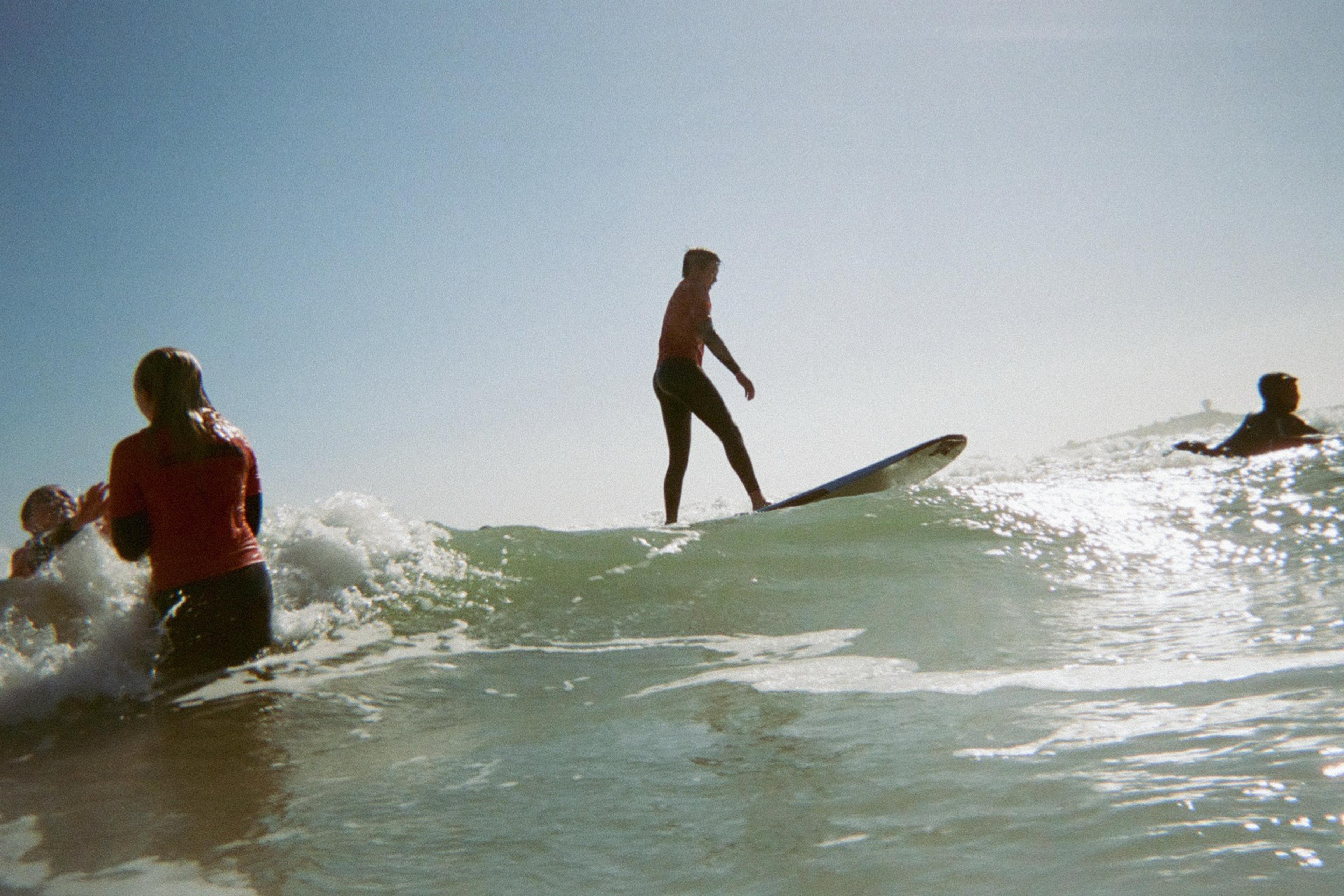 A person in a wetsuit rides a wave on a surfboard, surrounded by others in red tops, in bright sunlight and a clear sky.
