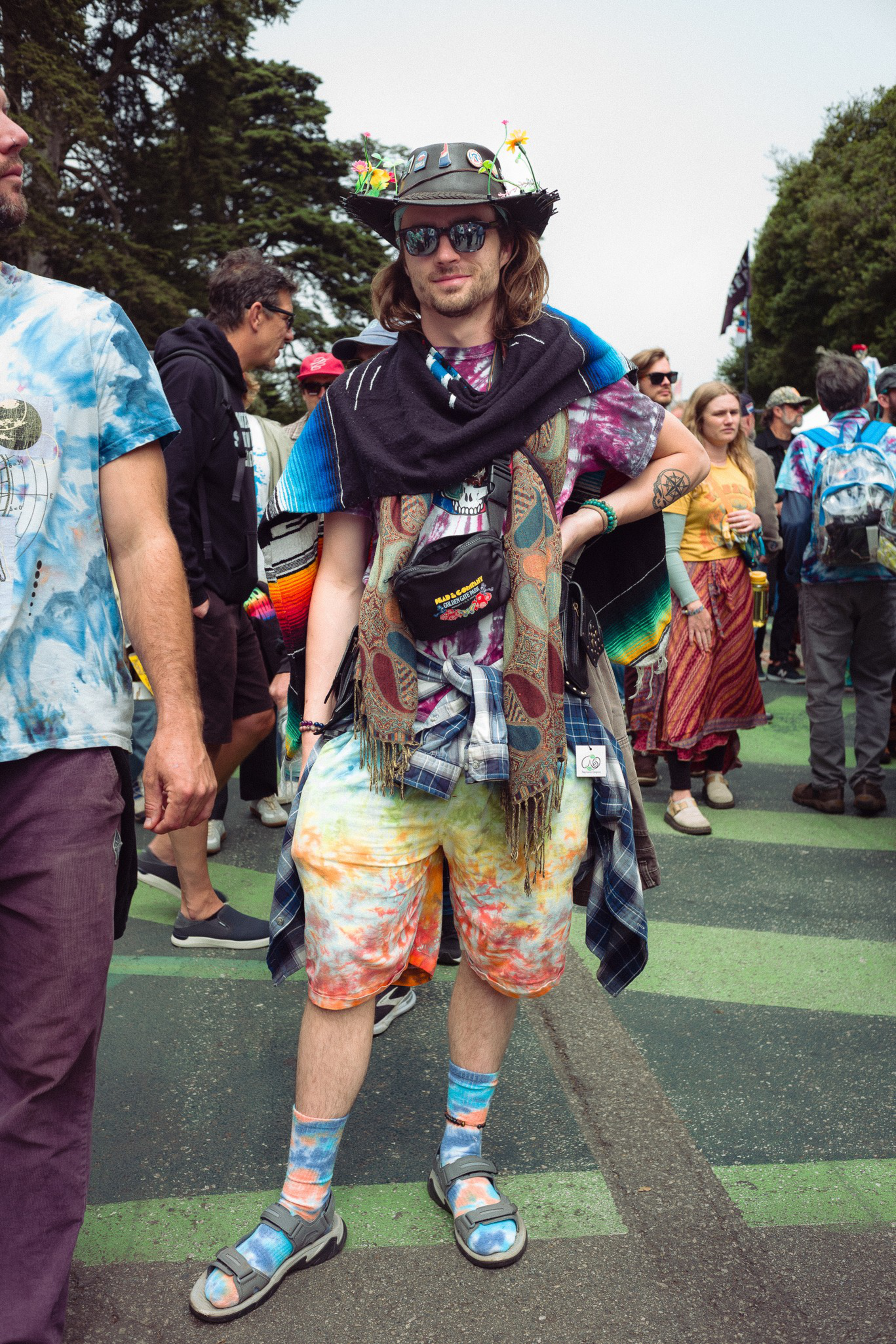 A person wearing a decorated black hat, sunglasses, multi-patterned scarf, tie-dye shorts, blue socks, and sandals stands confidently among a crowd outdoors.