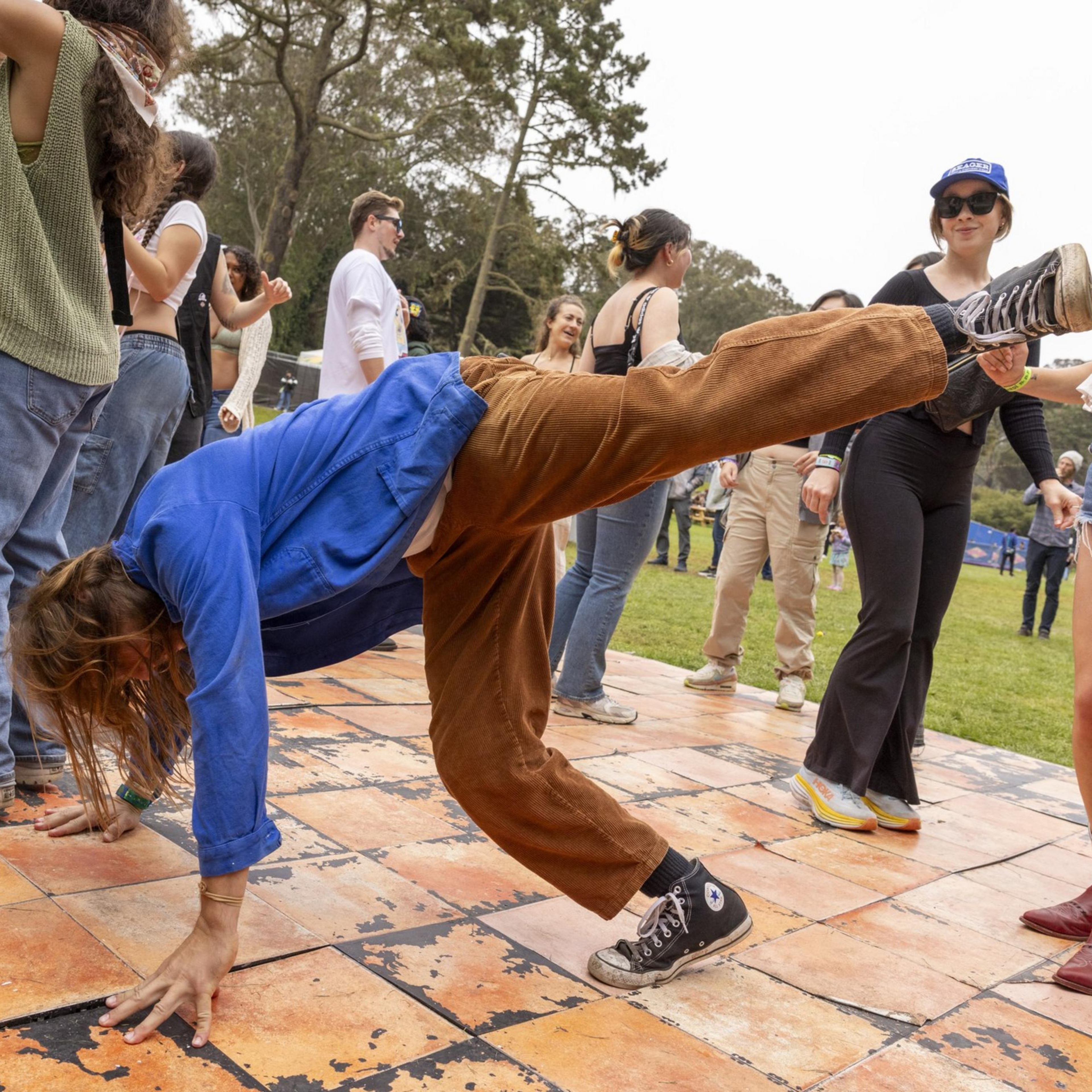 A person in a blue jacket and brown pants does a handstand on an outdoor dance floor while a group of people nearby dance and watch.