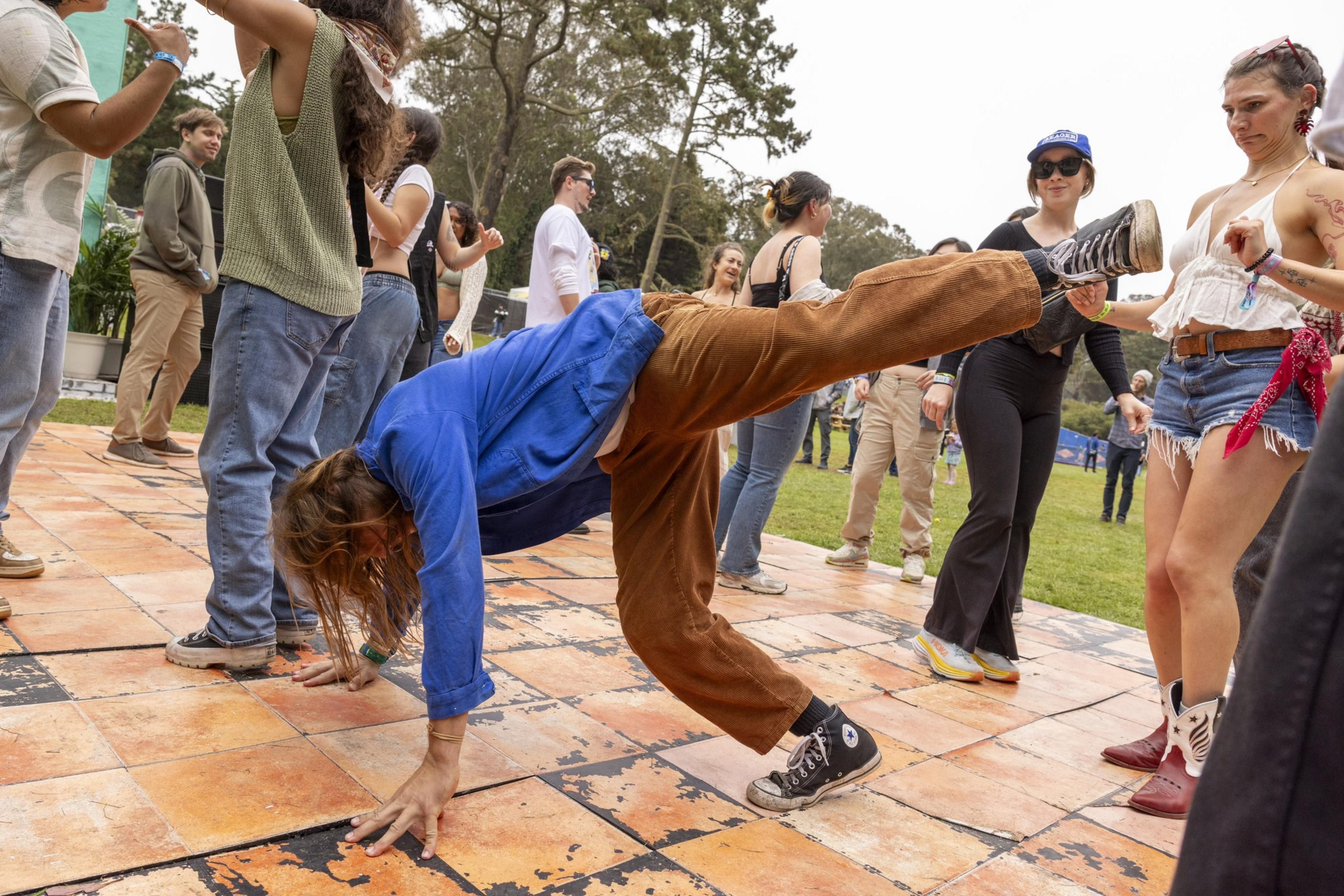 A person in a blue jacket and brown pants does a handstand on an outdoor dance floor while a group of people nearby dance and watch.
