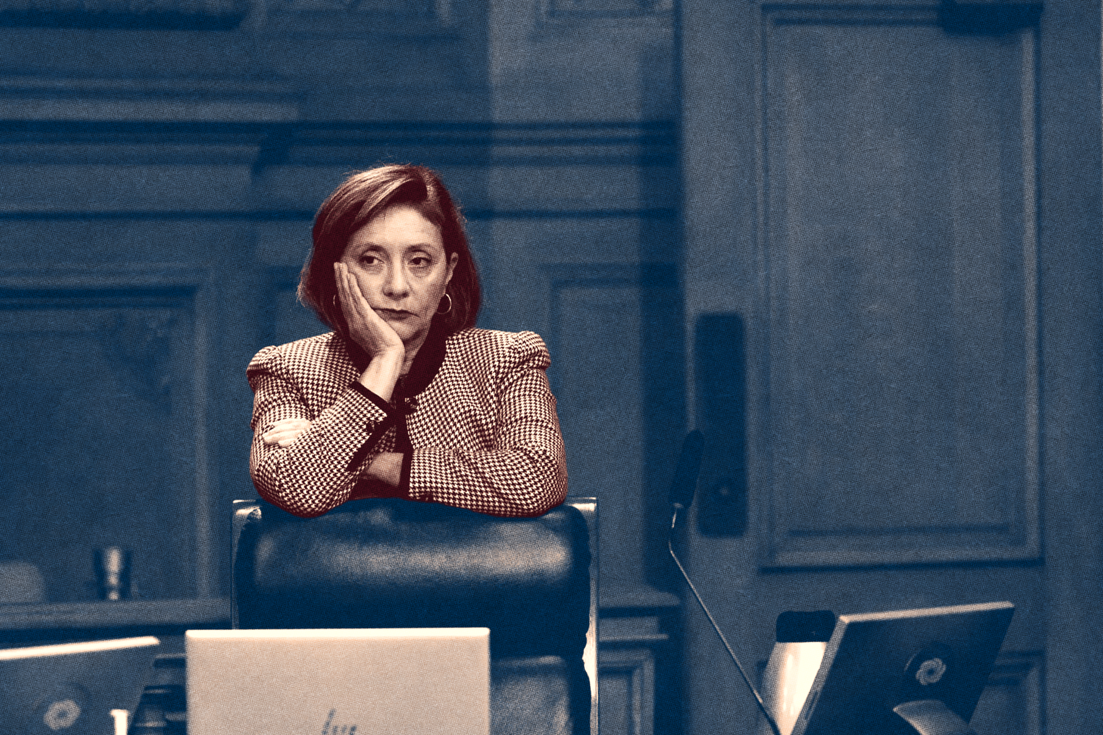 A woman with a pensive expression rests her head on her hand behind a desk with laptops and a microphone, in a formal room with paneled walls.