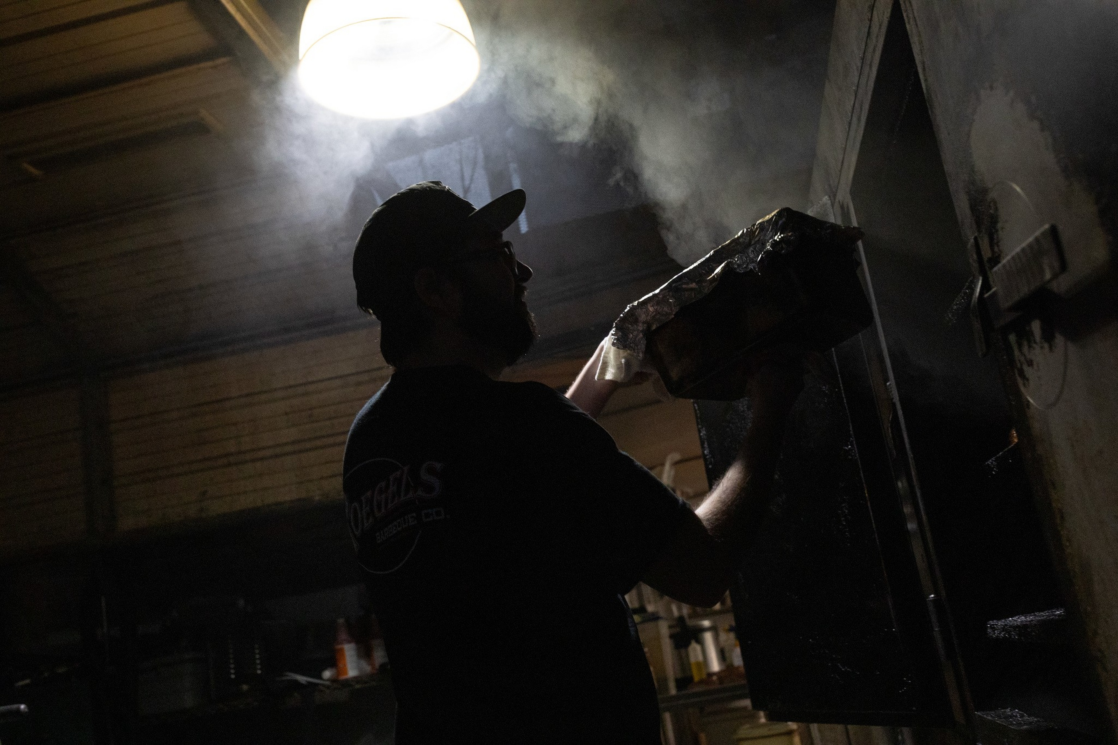 A man wearing a cap and glasses works in a smoky, dimly lit room, holding a tray wrapped in foil near a large, open metal oven or smoker.
