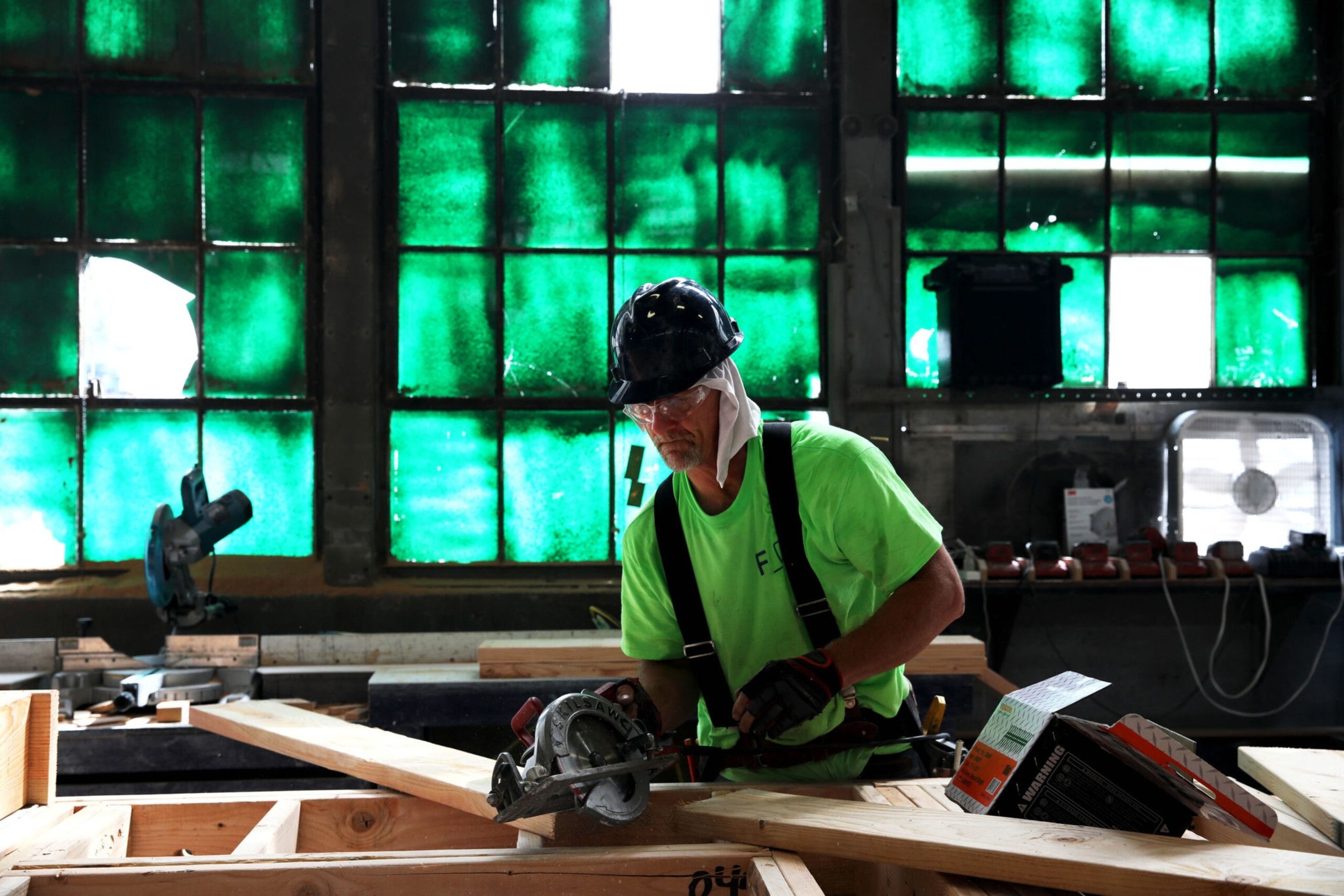 A construction worker wearing a green shirt, black hard hat, and gloves uses a circular saw to cut wood inside a workshop with green-tinted windows.