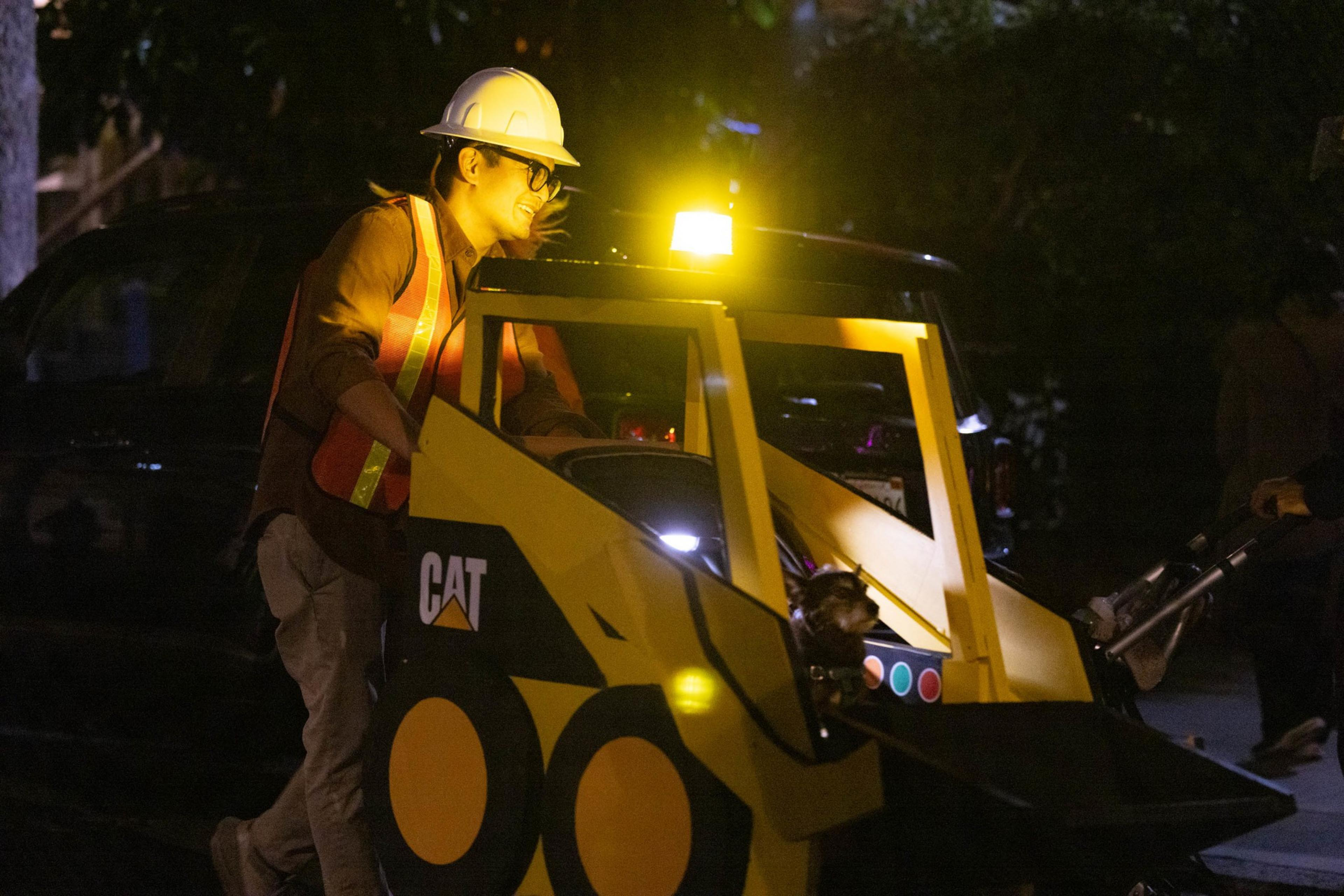 A person wearing a hard hat and safety vest operates a small, yellow CAT construction vehicle at night with a bright yellow light on top.