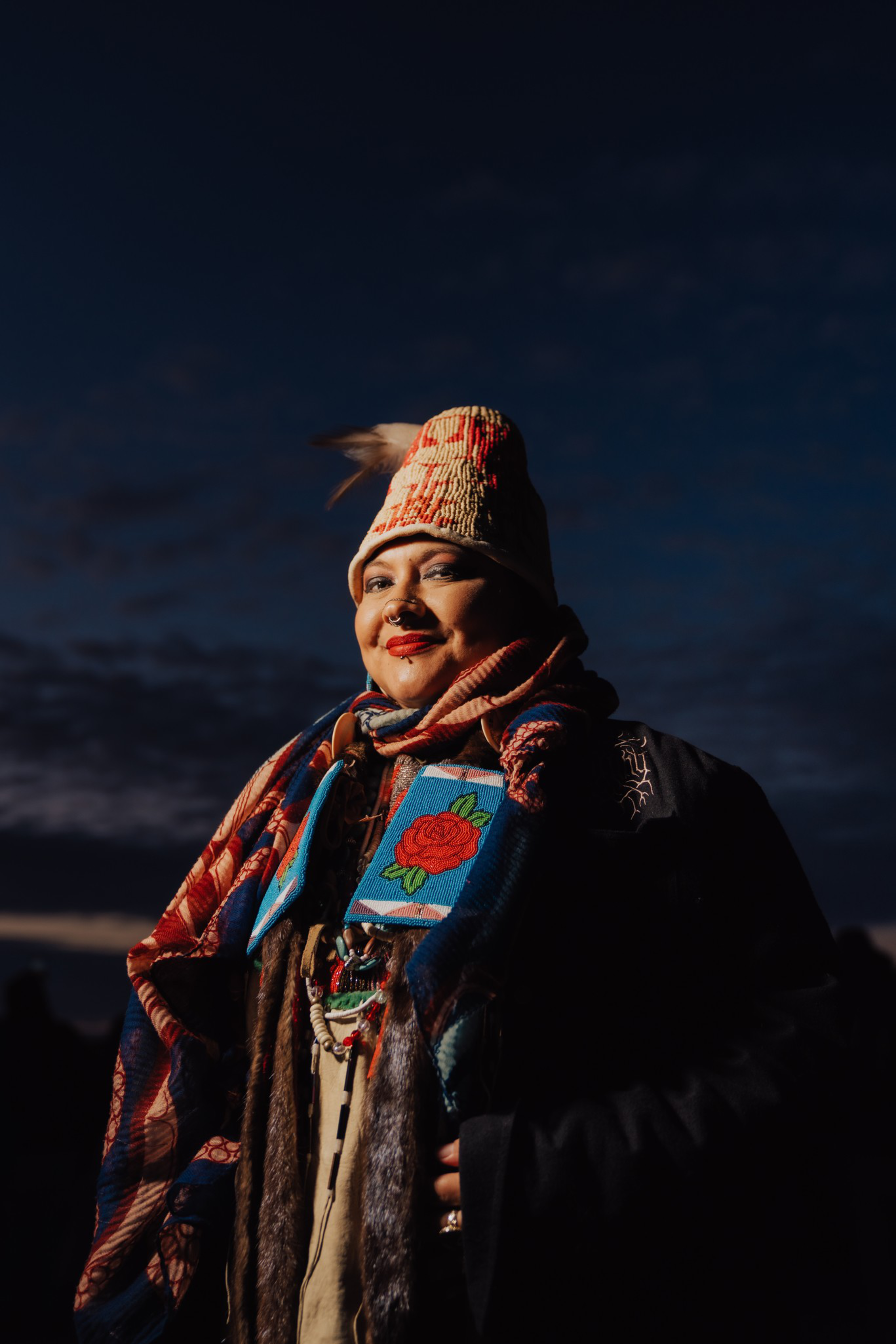 A person wearing a colorful woven hat, patterned scarf, and a beaded garment with a red rose design stands against a dark, cloudy sky.
