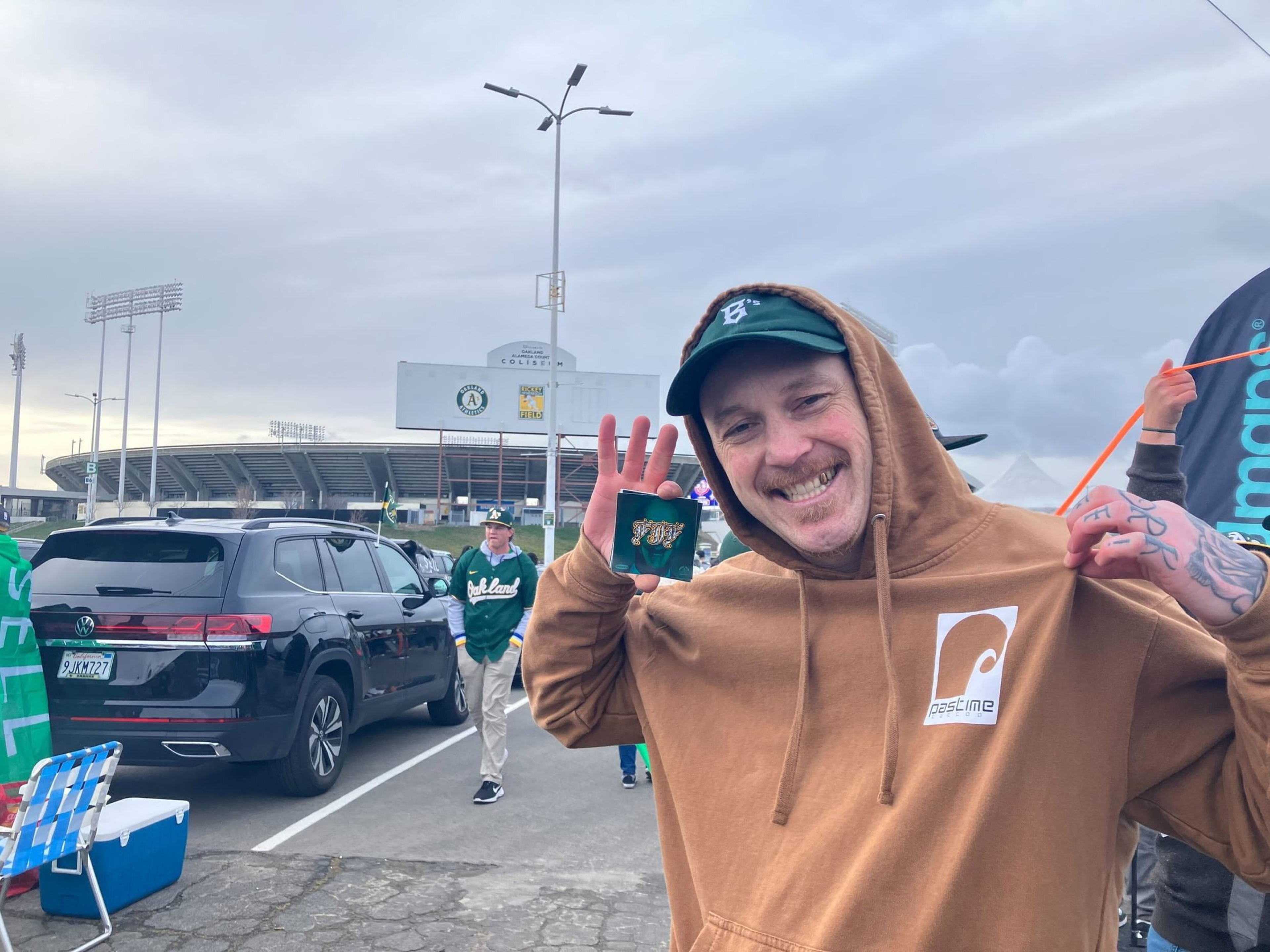 A man in a brown hoodie smiling, making a peace sign, with a stadium and fans in the background.