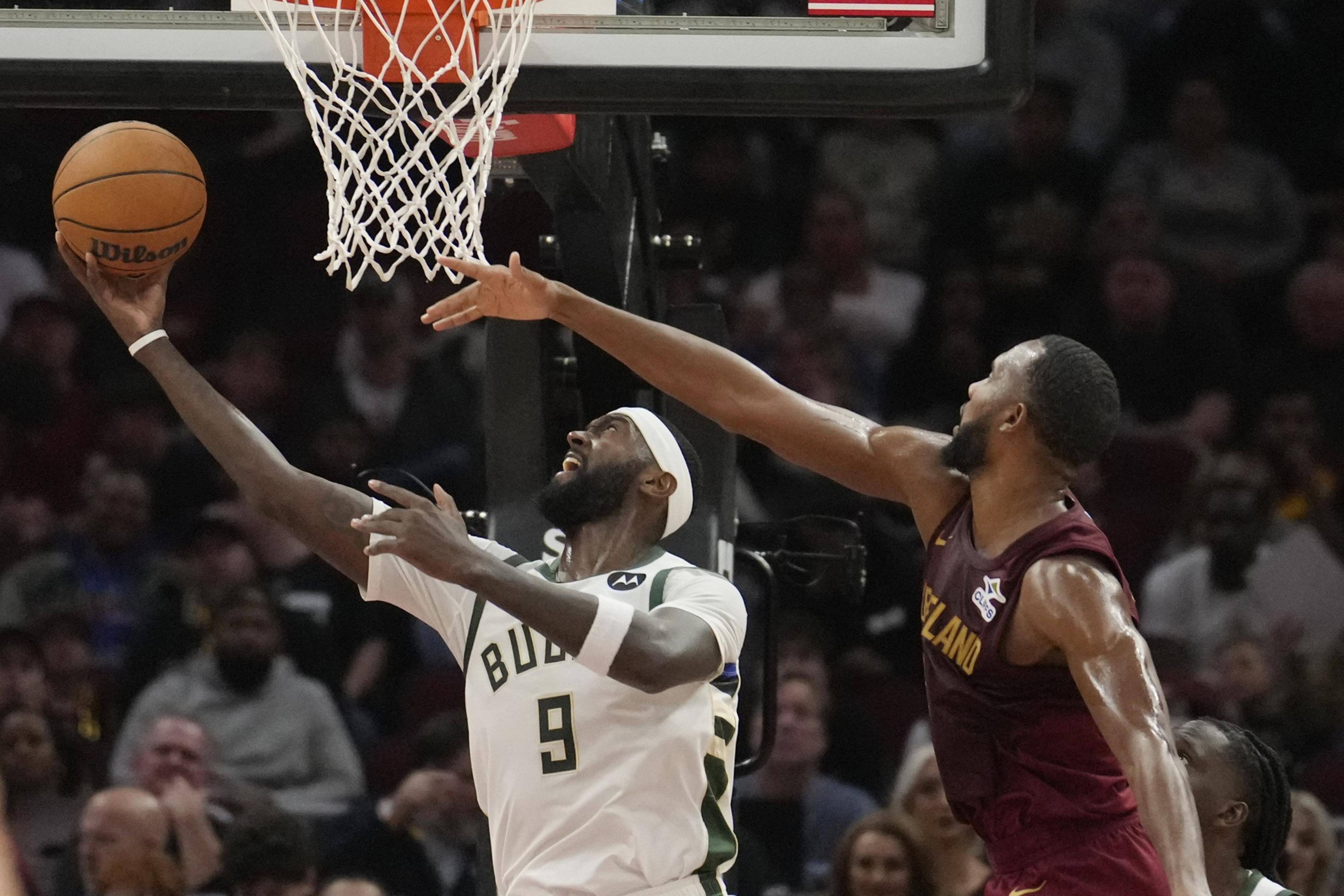 A basketball player in a white jersey attempts a layup while another player in a maroon jersey tries to block the shot. The scene is intense and focused.