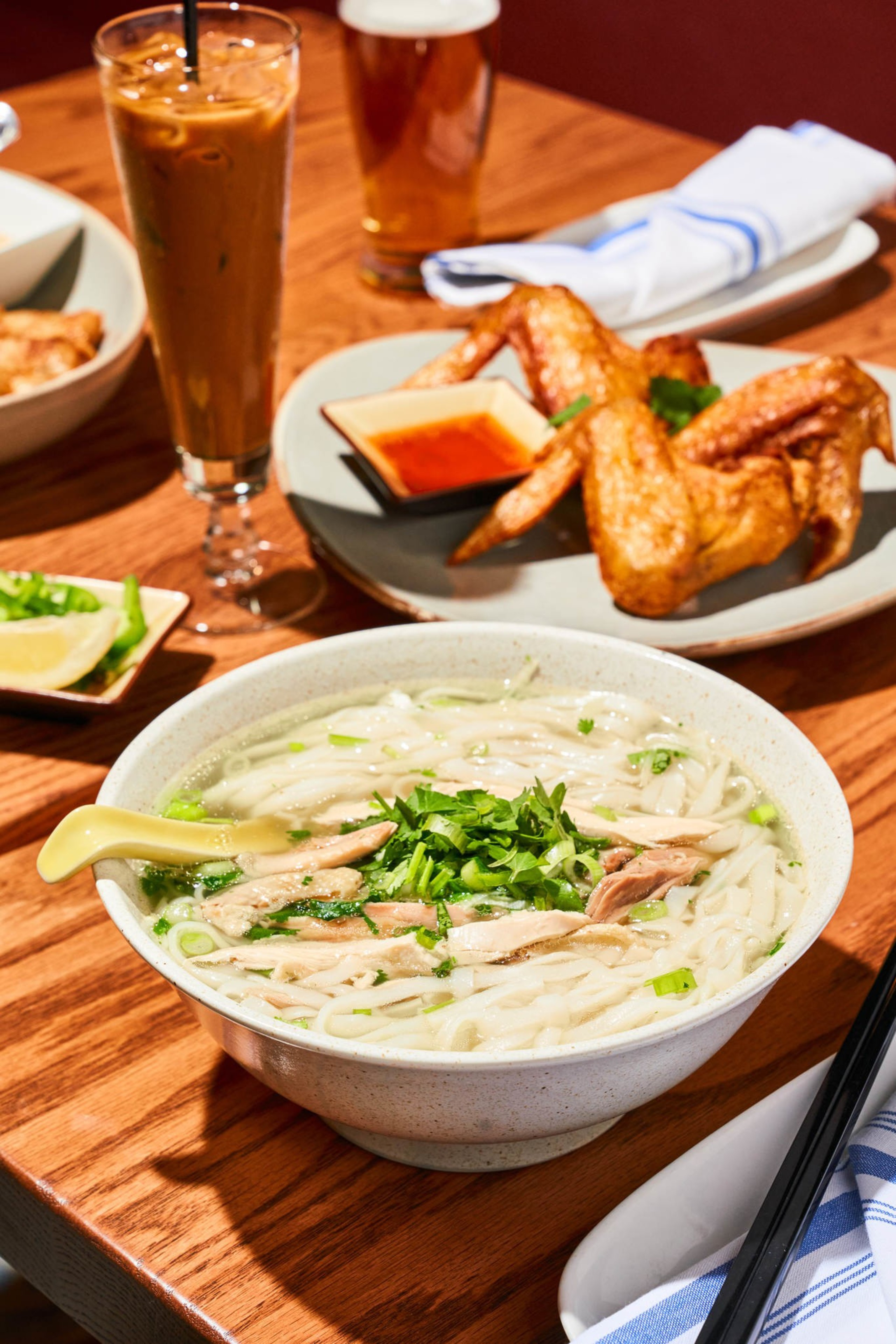 A bowl of noodle soup with herbs and meat, a plate of fried chicken wings with sauce, a glass of iced coffee, and a beverage glass are on a wooden table.