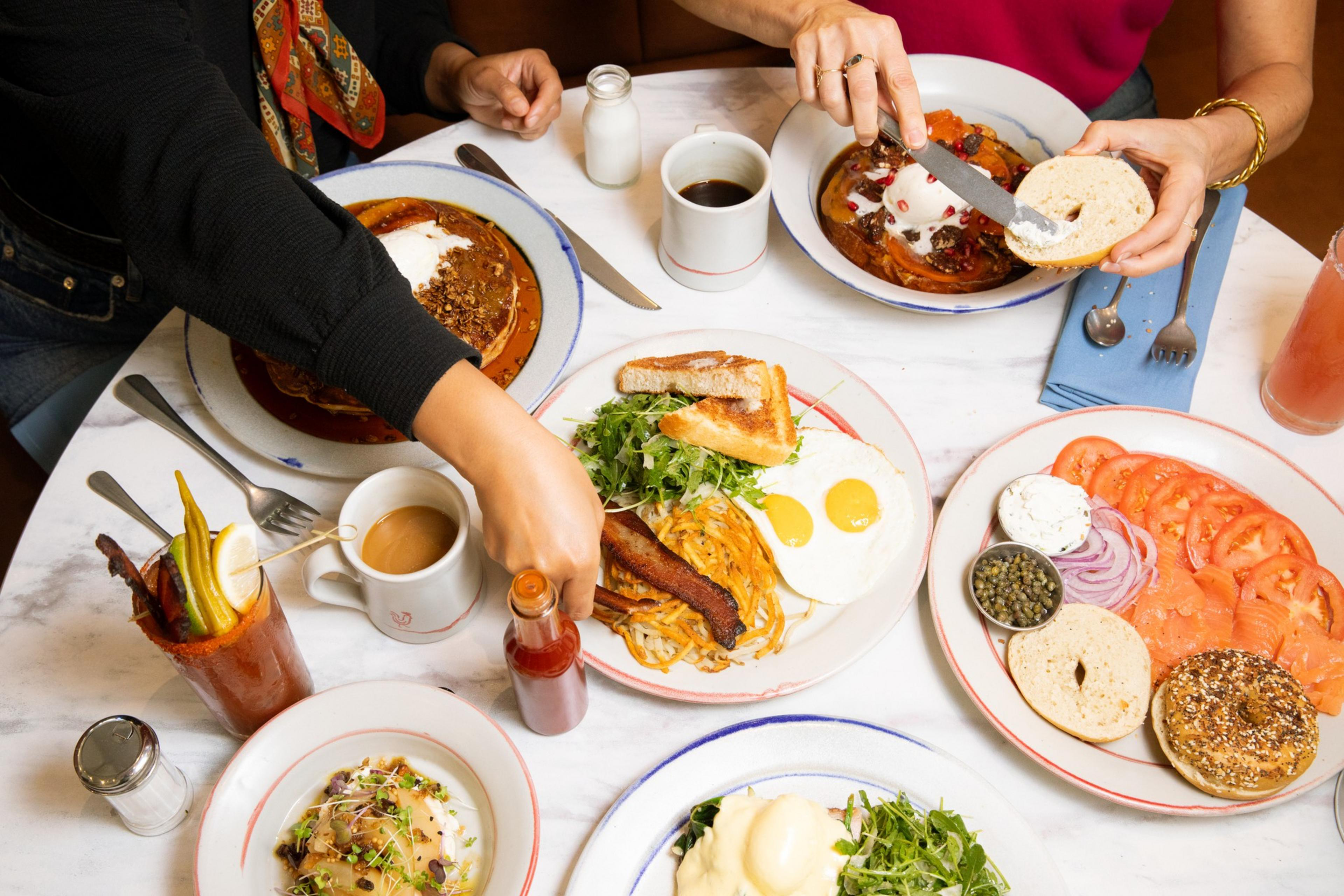 A breakfast table with various dishes, including pancakes, eggs, bacon, bagels, smoked salmon, and drinks. Two people are preparing their meals.