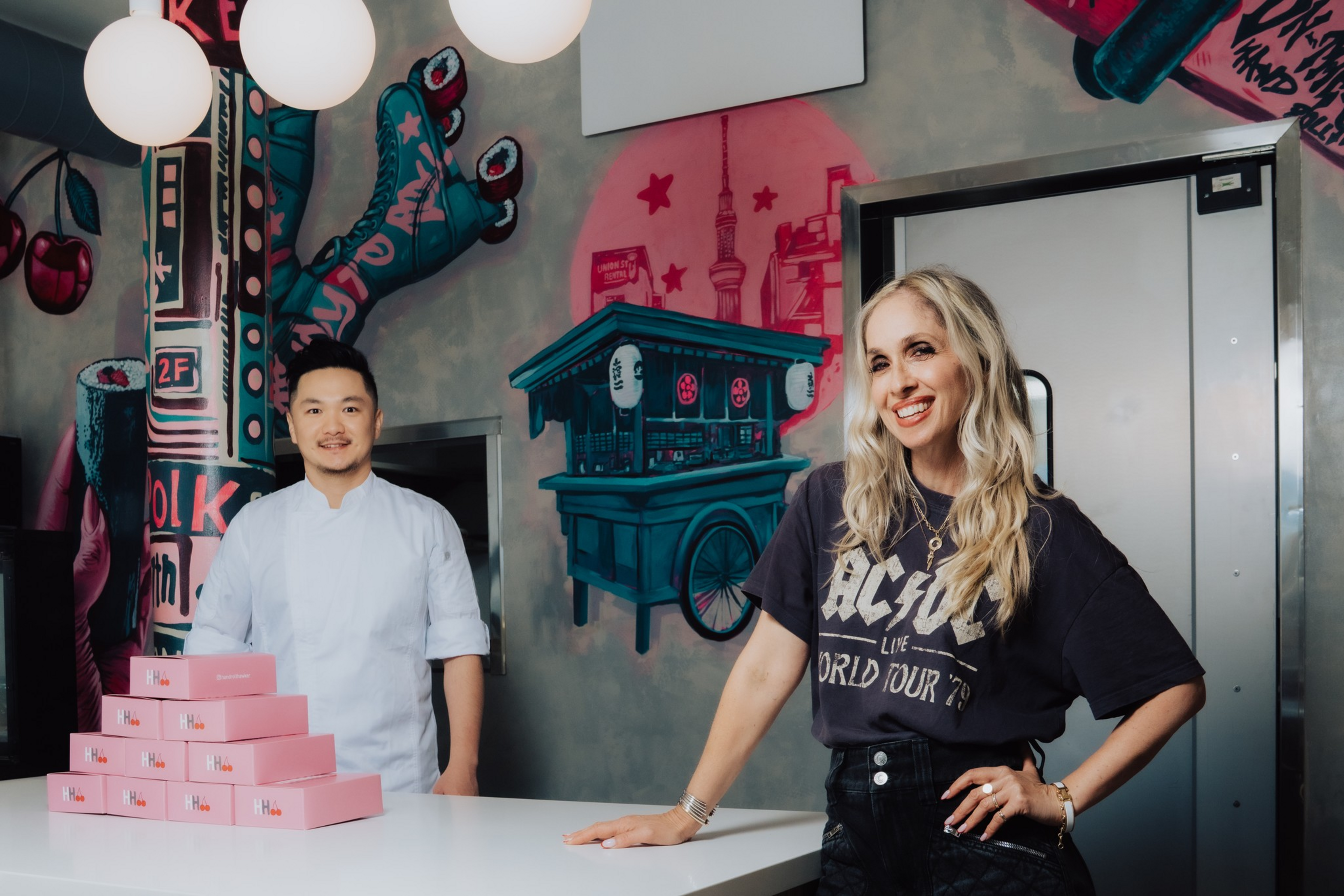 A man in a white chef coat stands behind a stack of pink boxes, while a smiling woman in a black AC/DC shirt leans on a counter in a colorful muraled room.