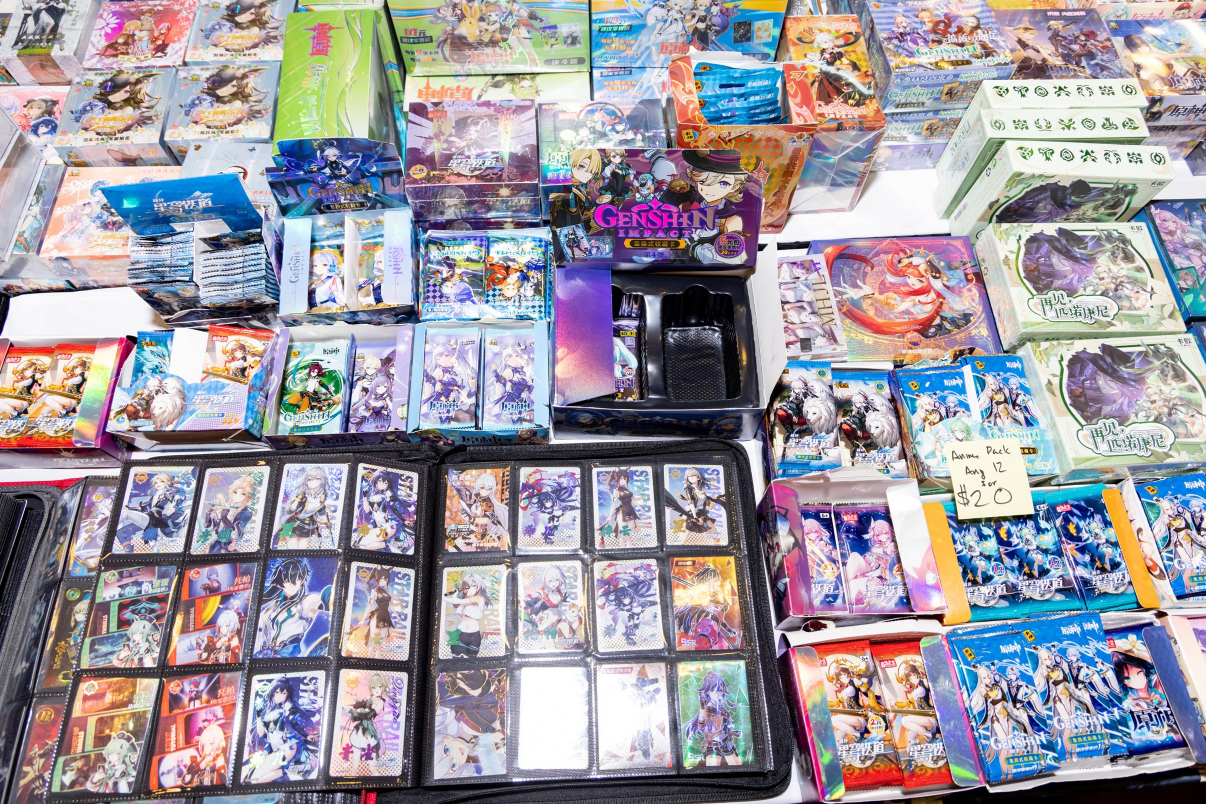 The image shows a display of various collectible trading cards and boxes, prominently featuring &quot;Genshin&quot; themed items, arranged on a table with some prices visible.