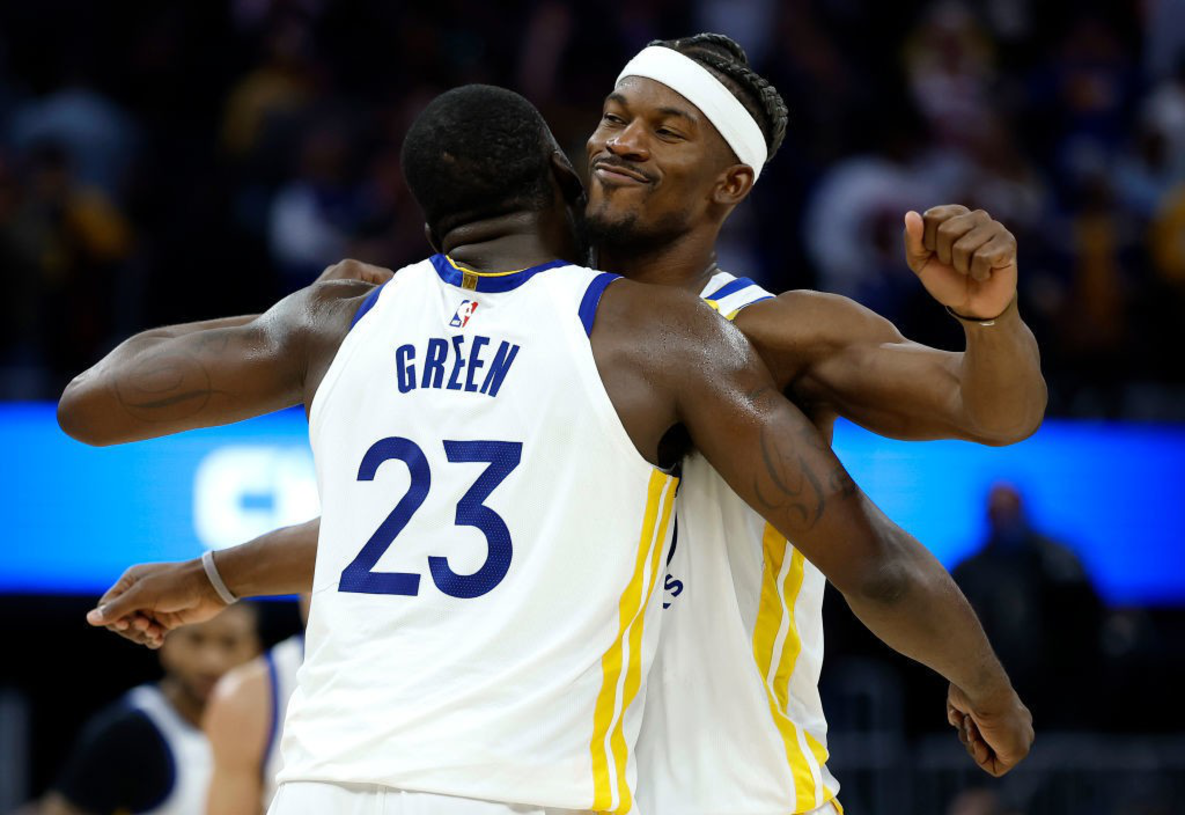 Two basketball players in white jerseys embrace with smiles, one wearing a headband and the other with "Green 23" on his back, against a blurred arena background.