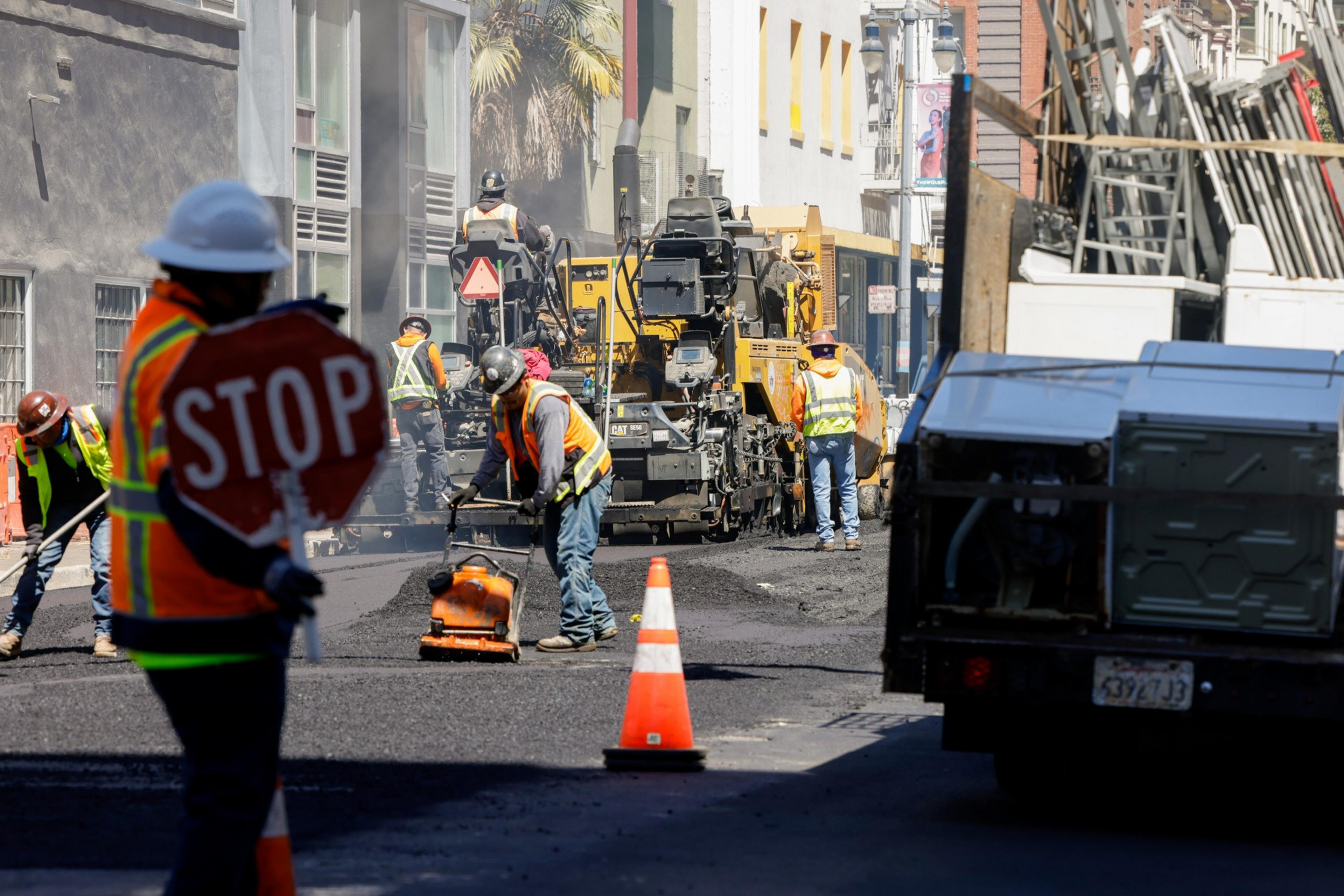 Construction workers in safety vests and helmets are operating machinery and tools to pave a road. One worker holds a "STOP" sign, and orange traffic cones are placed nearby.