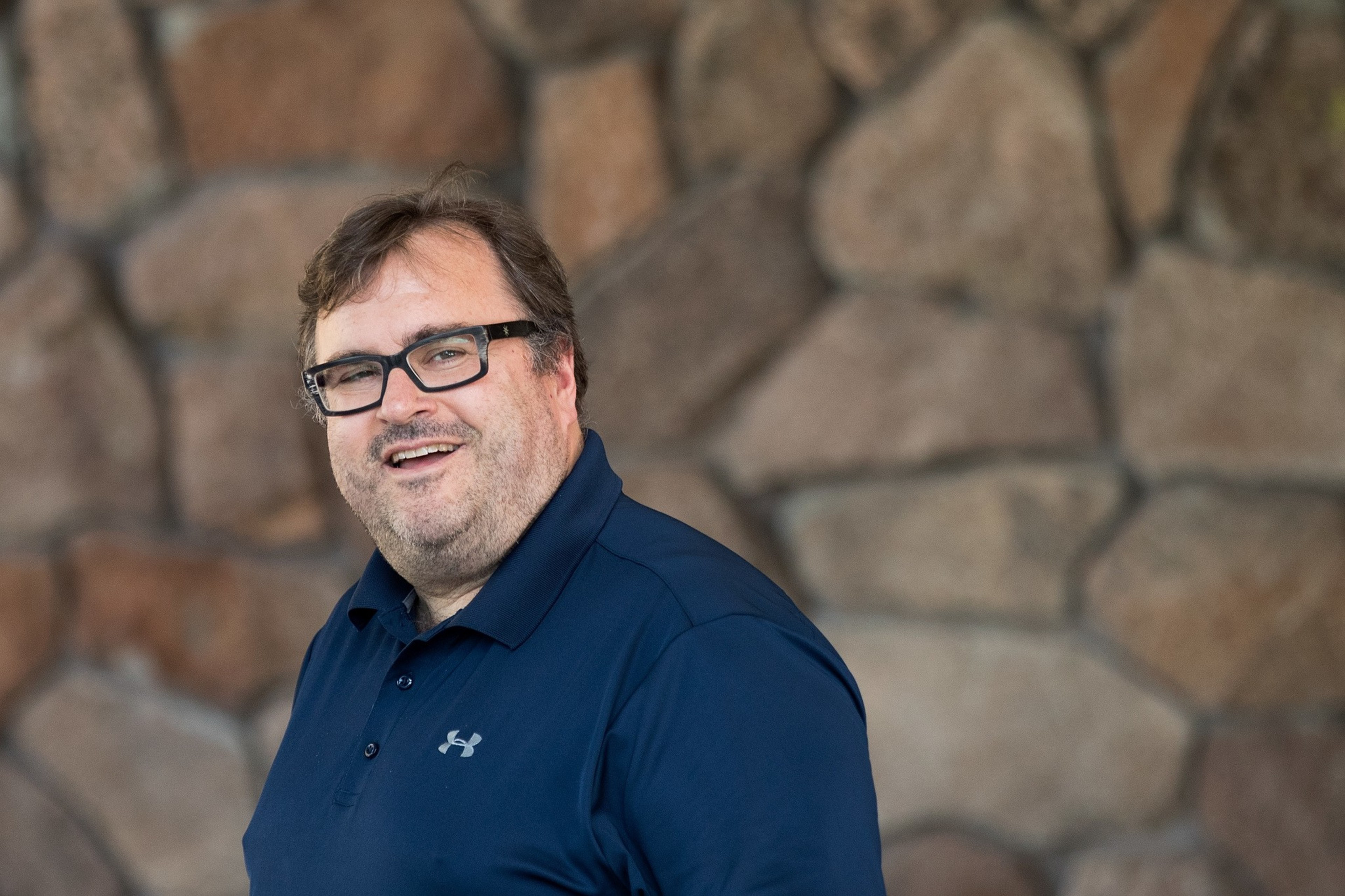 A smiling man with glasses and messy hair is wearing a navy blue Under Armour polo shirt. He stands in front of a stone wall background.