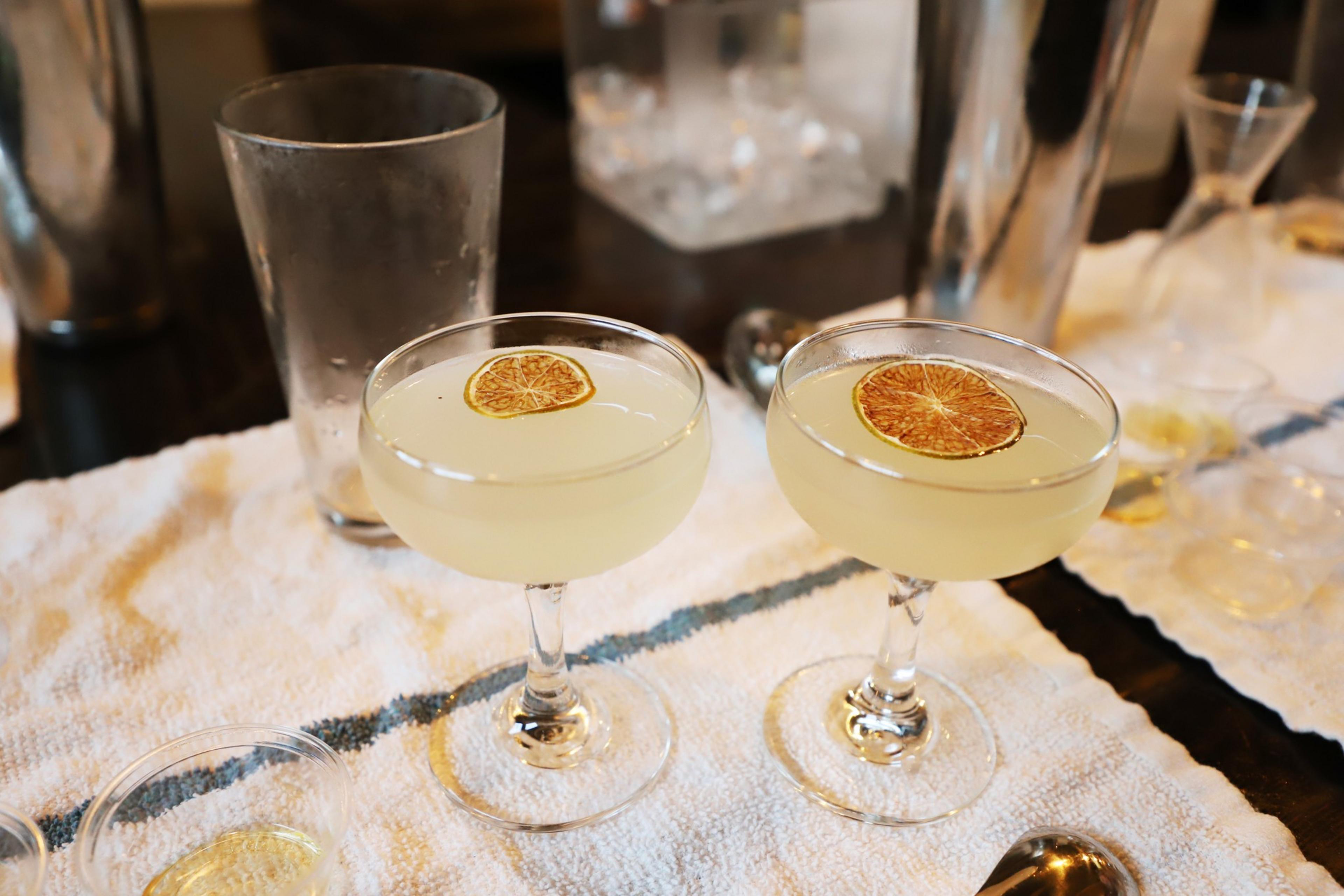 Two coupe glasses filled with a pale yellow drink are garnished with a dried citrus slice, set on a white cloth, with cocktail-making tools around.