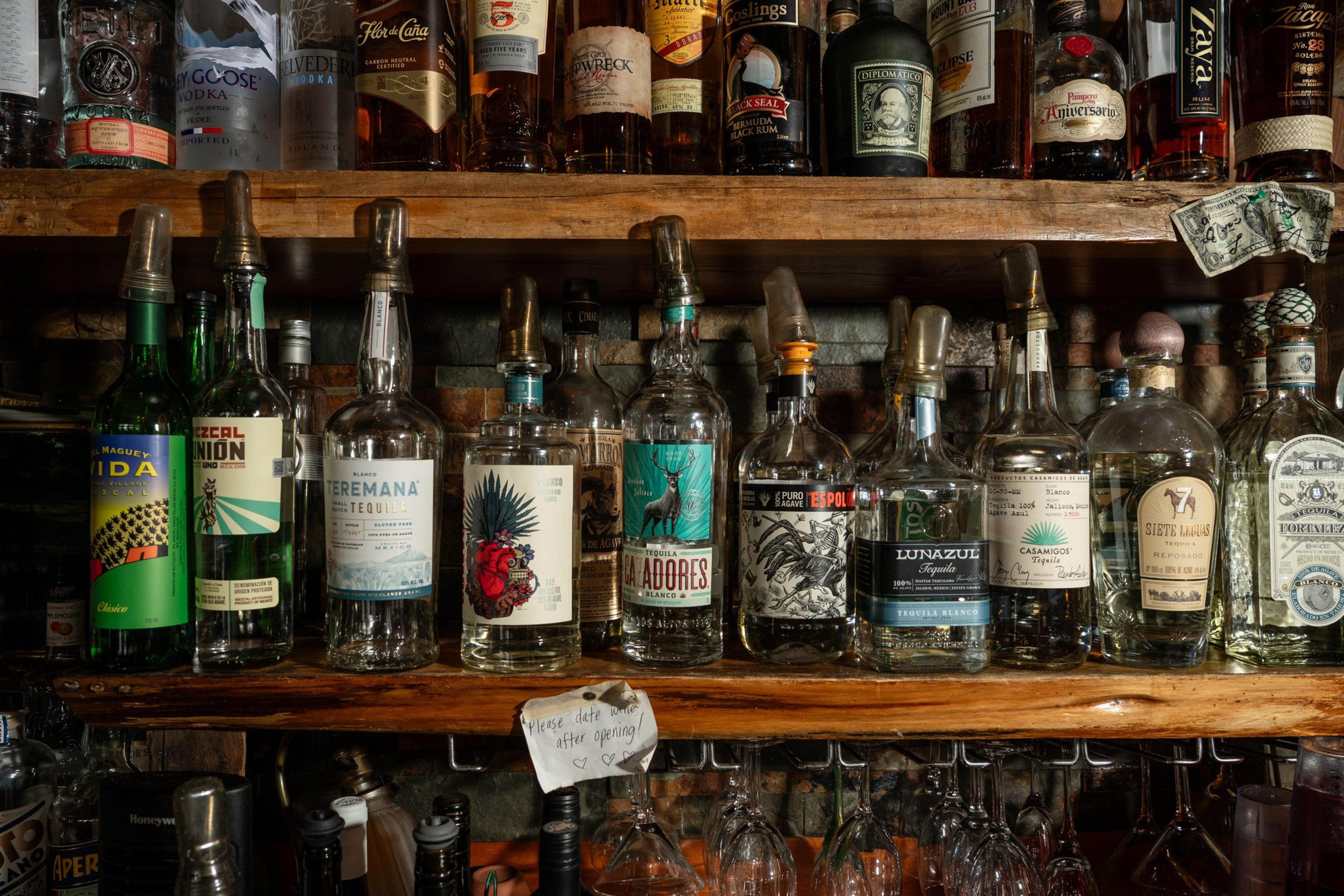 The image shows a shelf filled with various liquor bottles, mostly tequila and rum, with diverse labels. A small sign asks to date bottles after opening.
