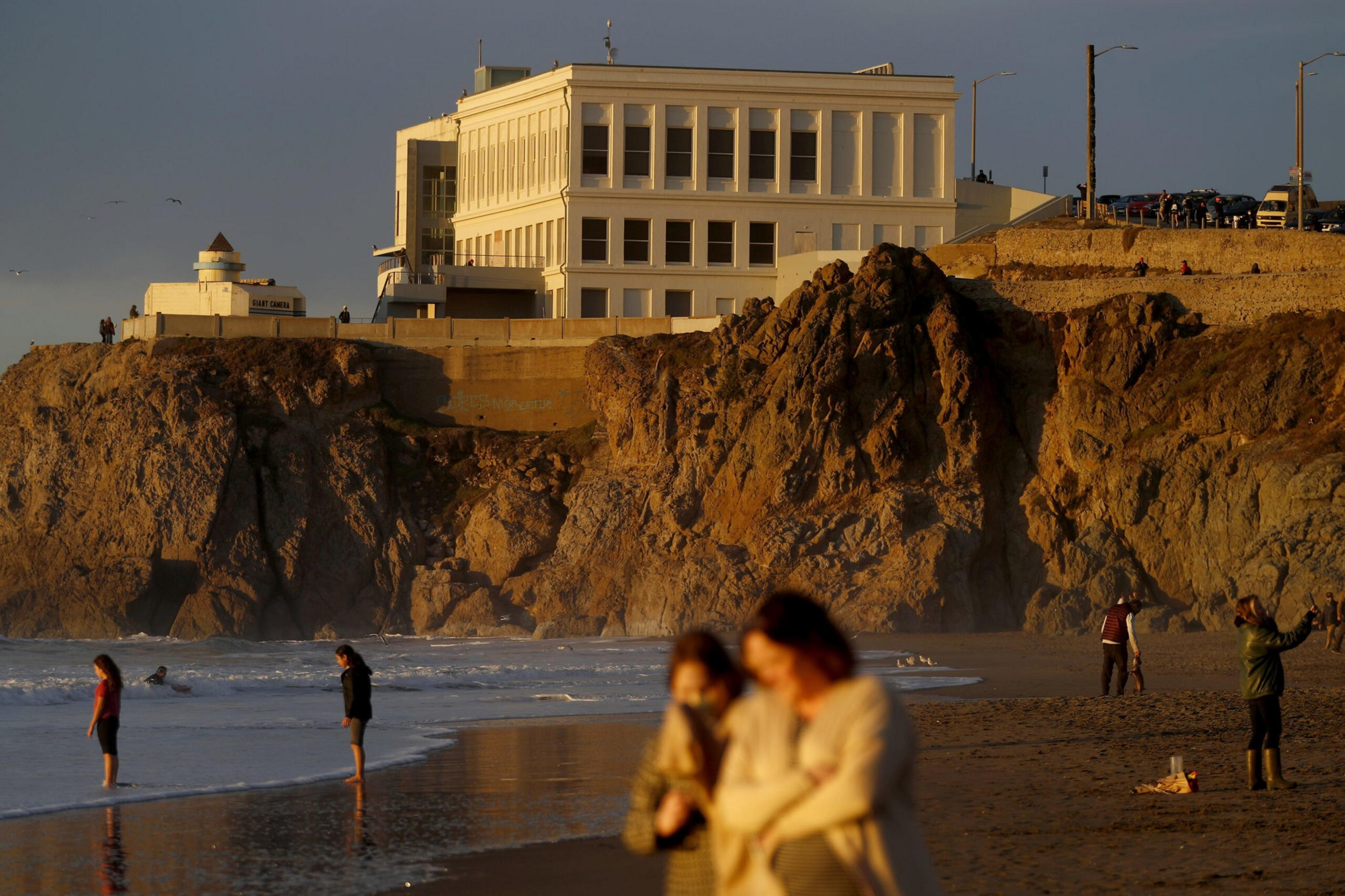 People walk and stand along a beach at sunset, with rocky cliffs and a large white building on top in the background.