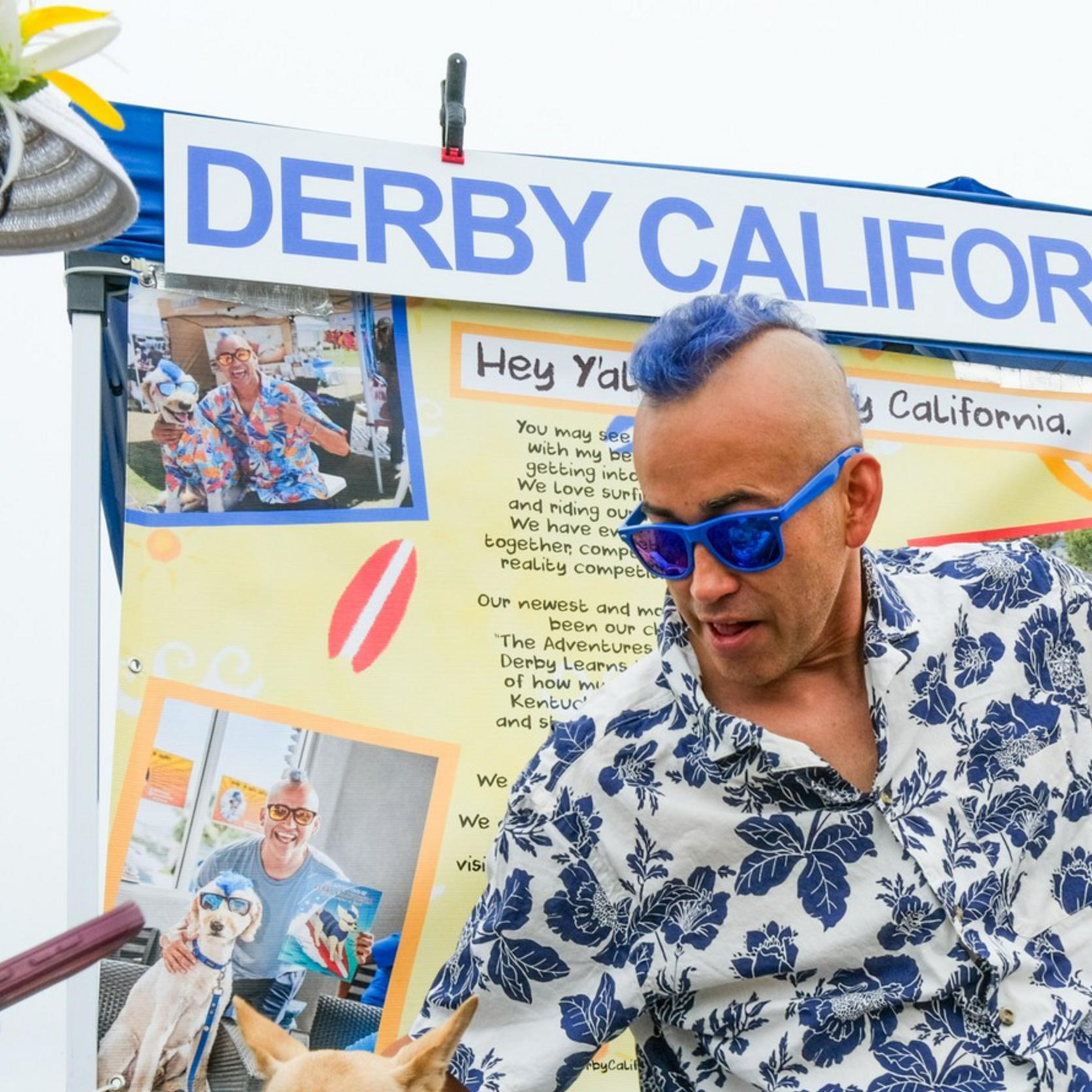 A man and two dogs, all wearing matching Hawaiian shirts, sit at a surf dog booth. One dog also wears sunglasses, while someone interacts with the smaller dog.