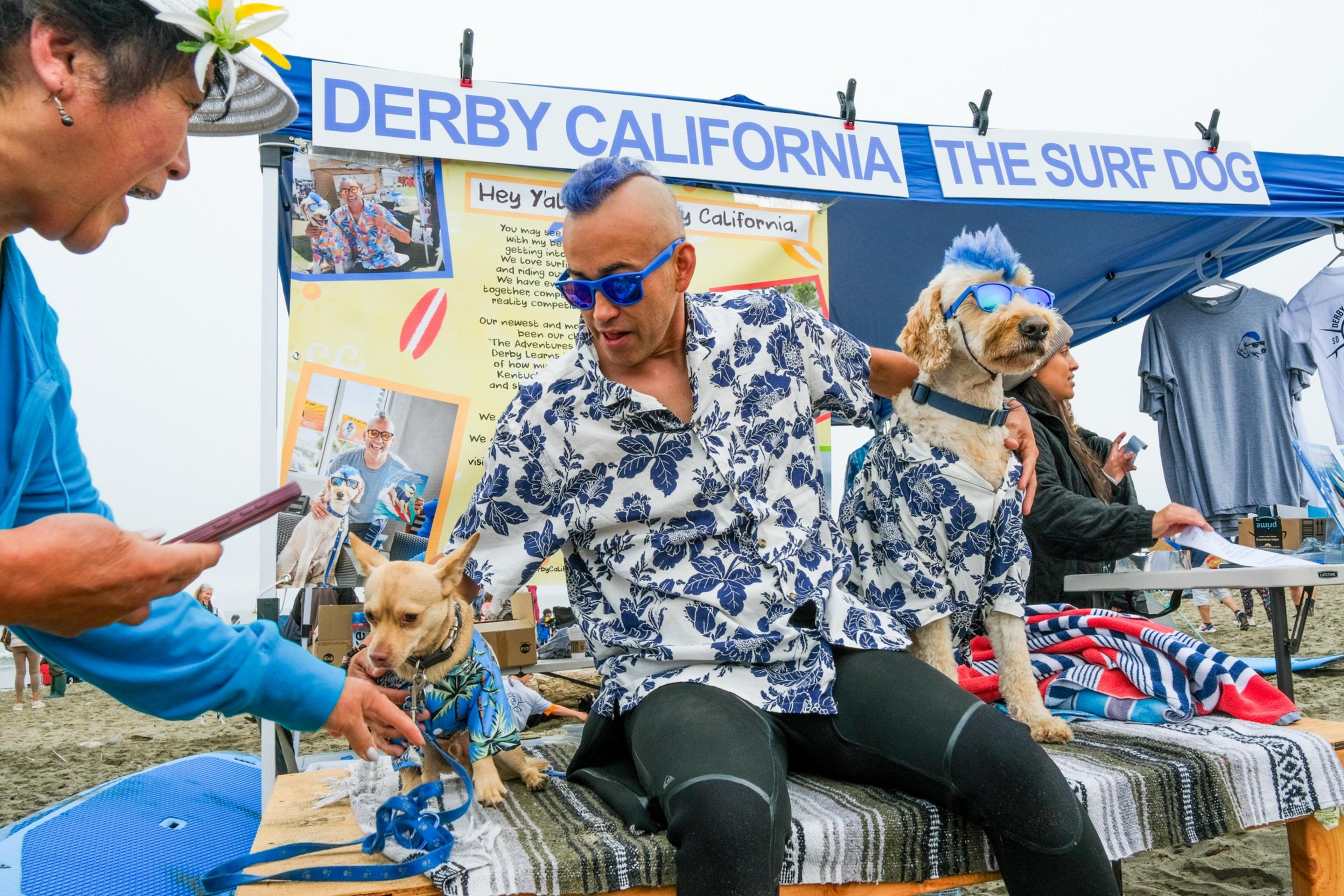 A man and two dogs, all wearing matching Hawaiian shirts, sit at a surf dog booth. One dog also wears sunglasses, while someone interacts with the smaller dog.