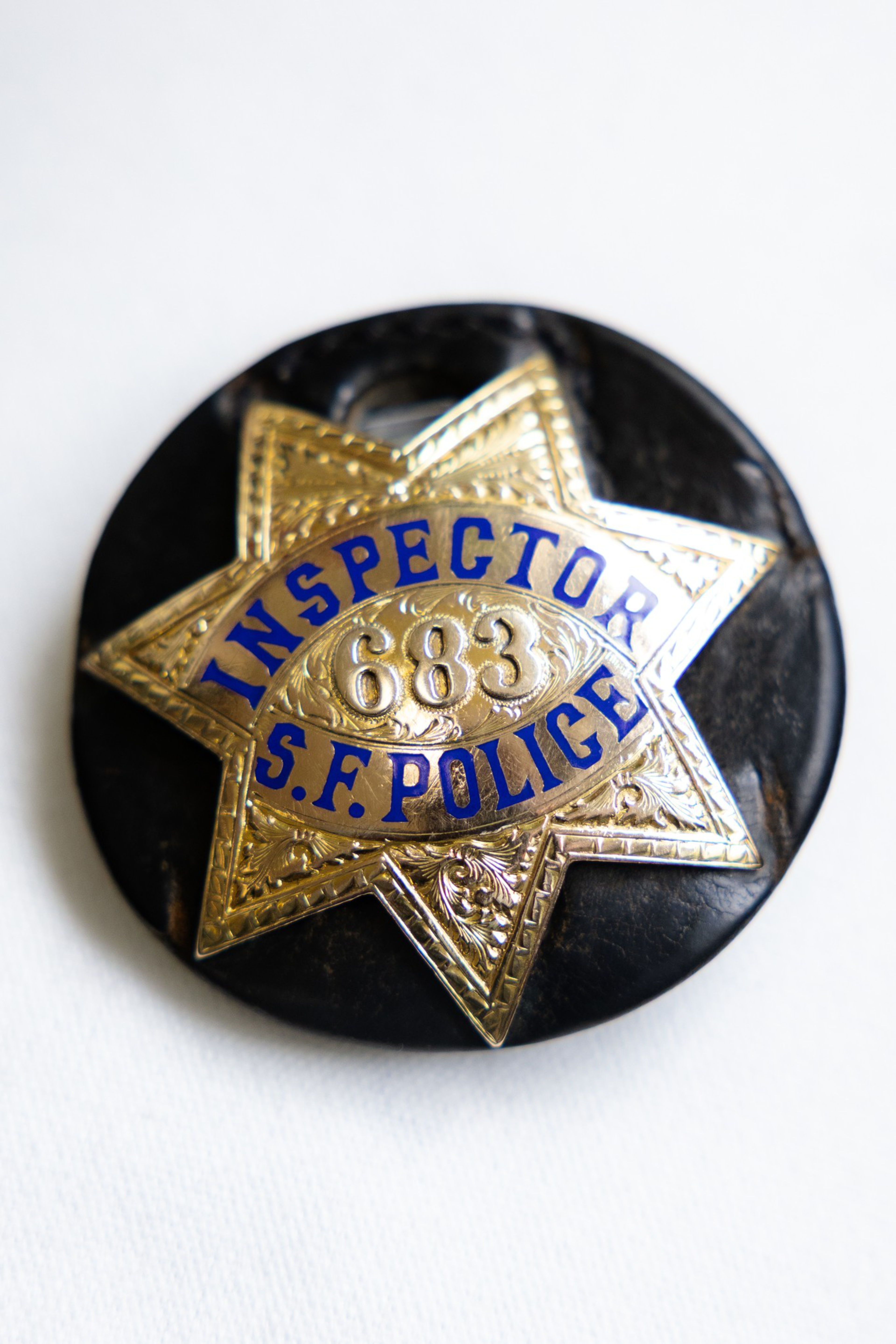 A gold star-shaped badge with “INSPECTOR,” “S.F. POLICE,” and the number “683" in blue letters, mounted on a round black backing.