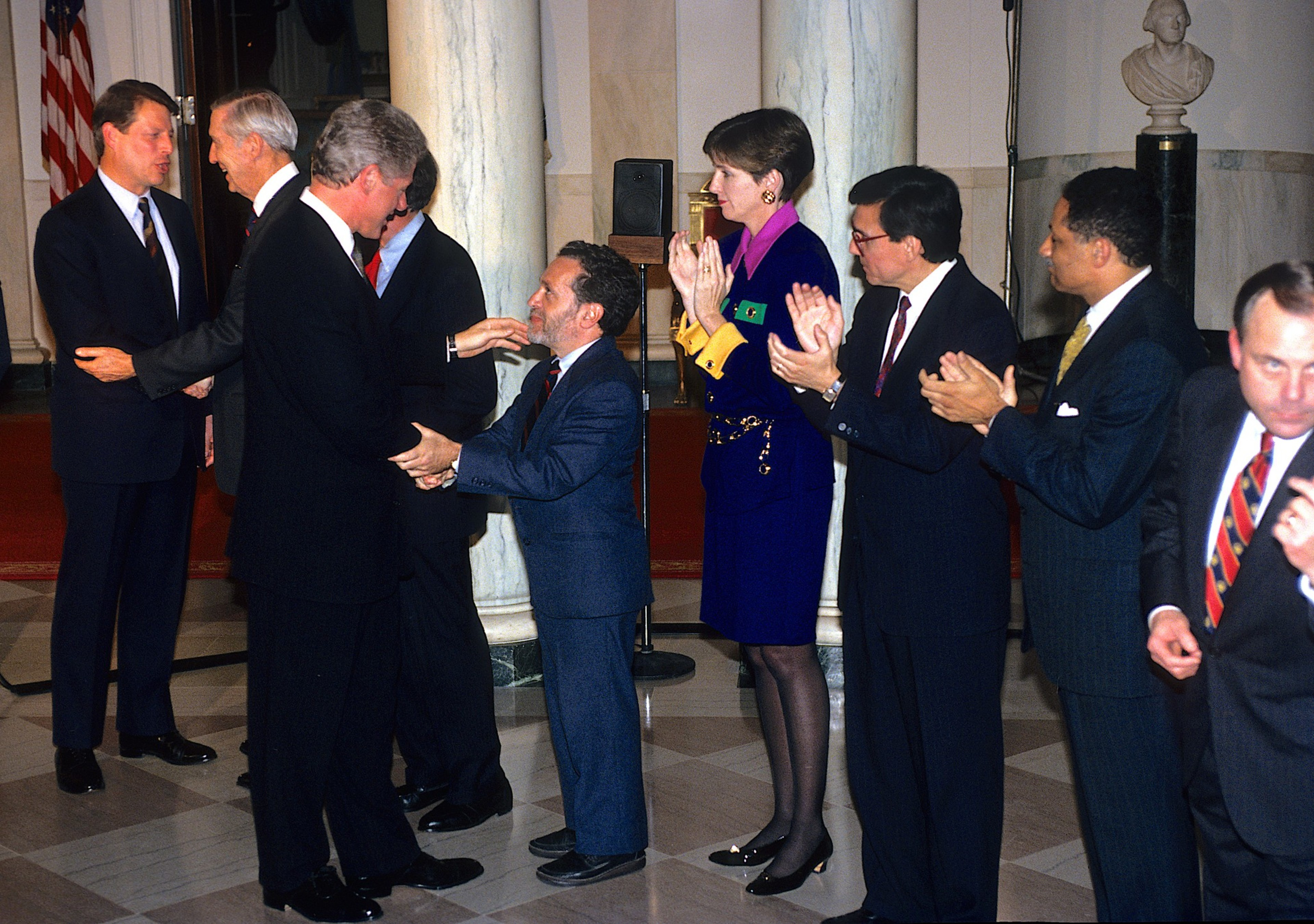A line of formally dressed people, with some shaking hands and others clapping, stands inside a marble-columned building with red carpet and statues.