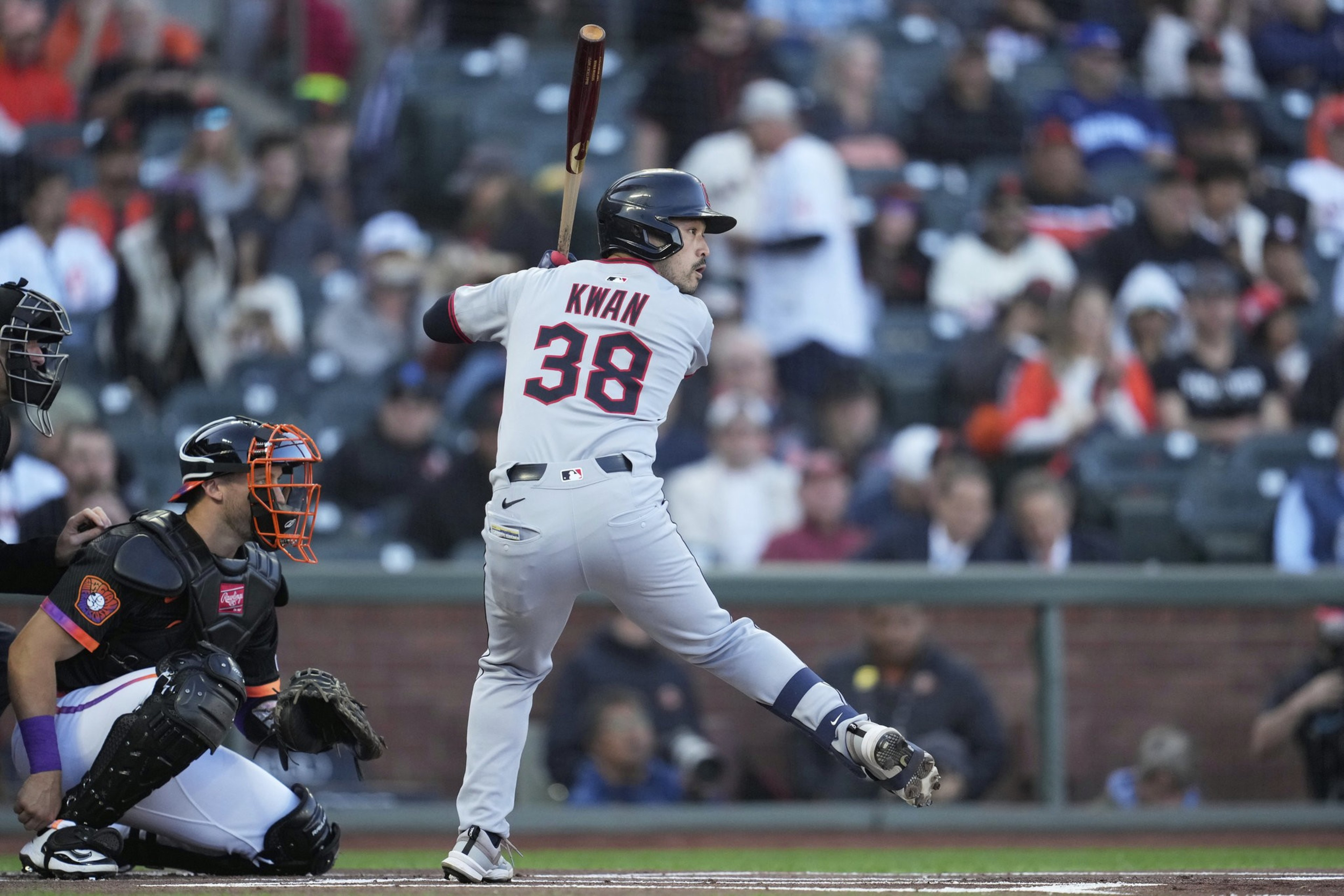 A baseball player, wearing a jersey with the number 38, swings a bat mid-action. A catcher and an umpire crouch nearby, with a blurred crowd in the background.