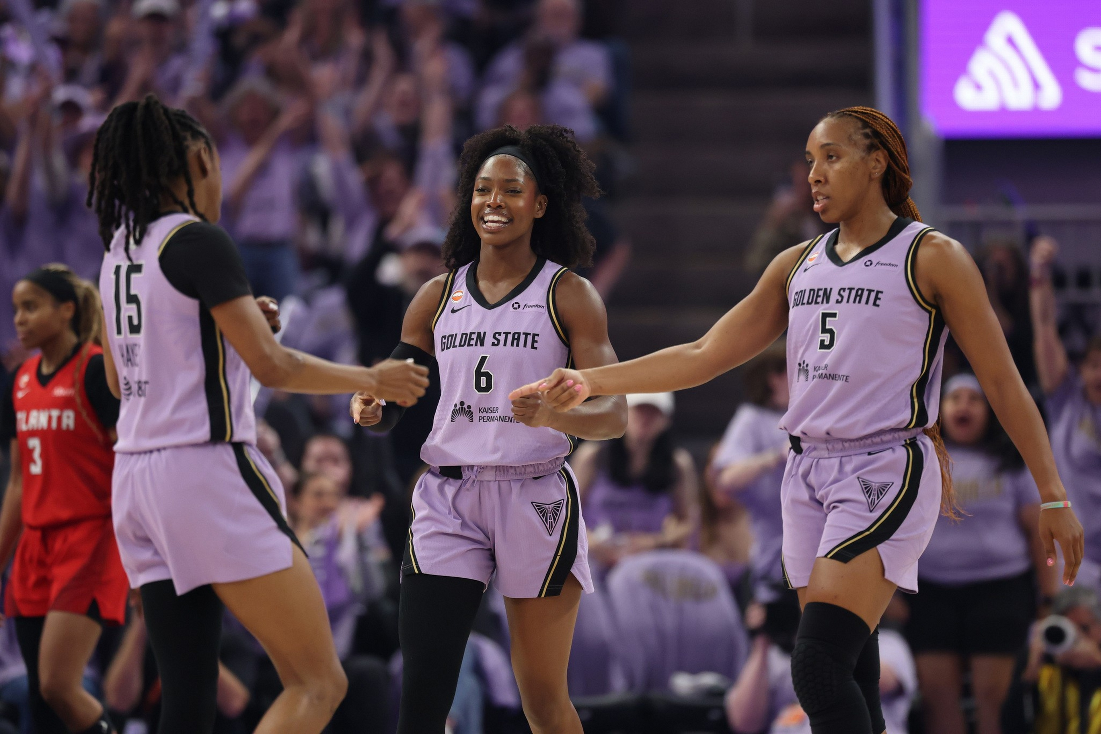Three female basketball players in Golden State uniforms celebrate on the court, with one wearing number 6 smiling between teammates number 15 and 5.