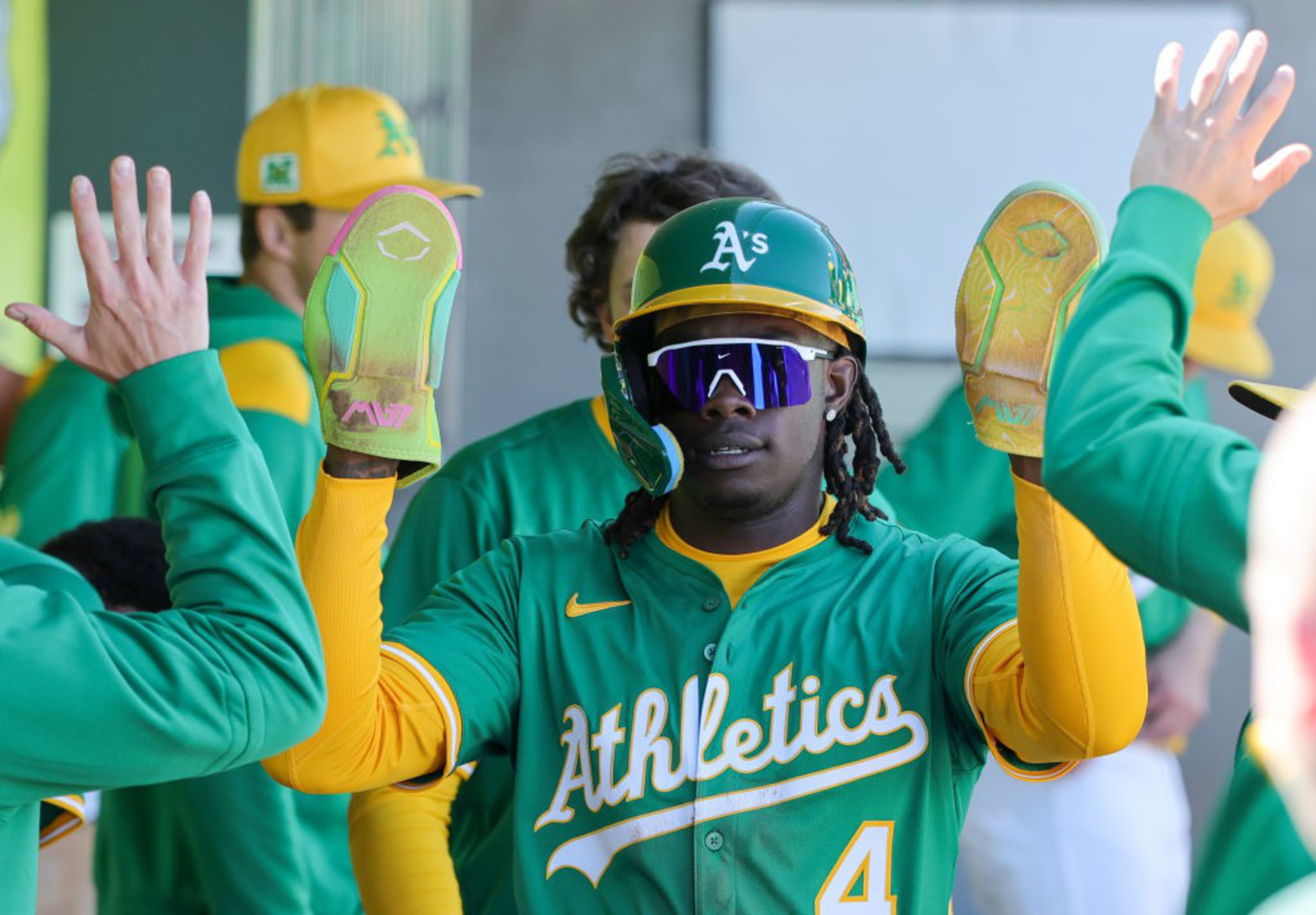 A baseball player in green and yellow gear high-fives teammates. He wears sunglasses and holds batting gloves. His team, &quot;Athletics,&quot; is visible on his jersey.