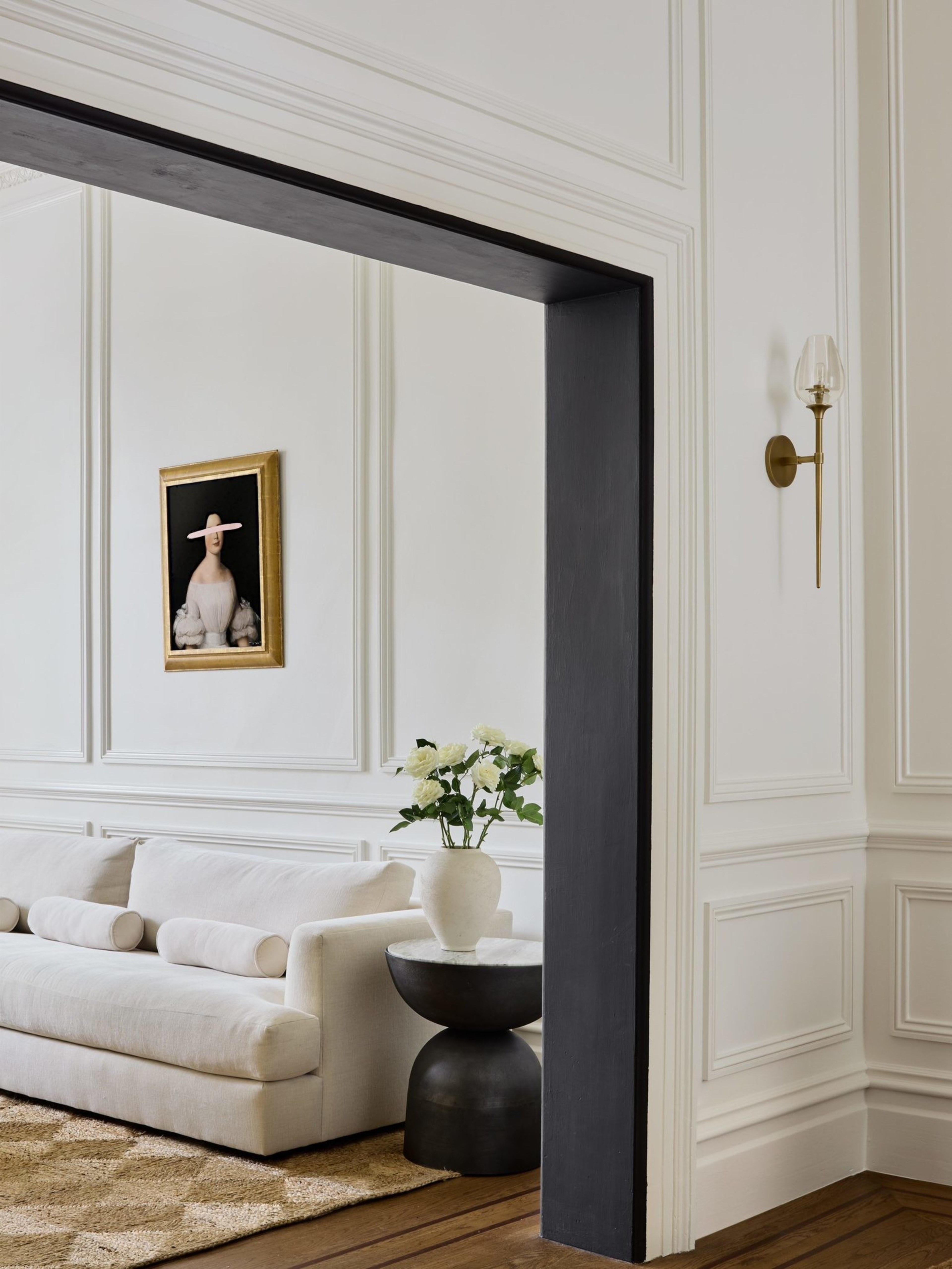 A stylish room features a white couch, a vase of white flowers on a black side table, a beige rug, and a wall painting of a person in a hat. Elegant paneled walls add sophistication.